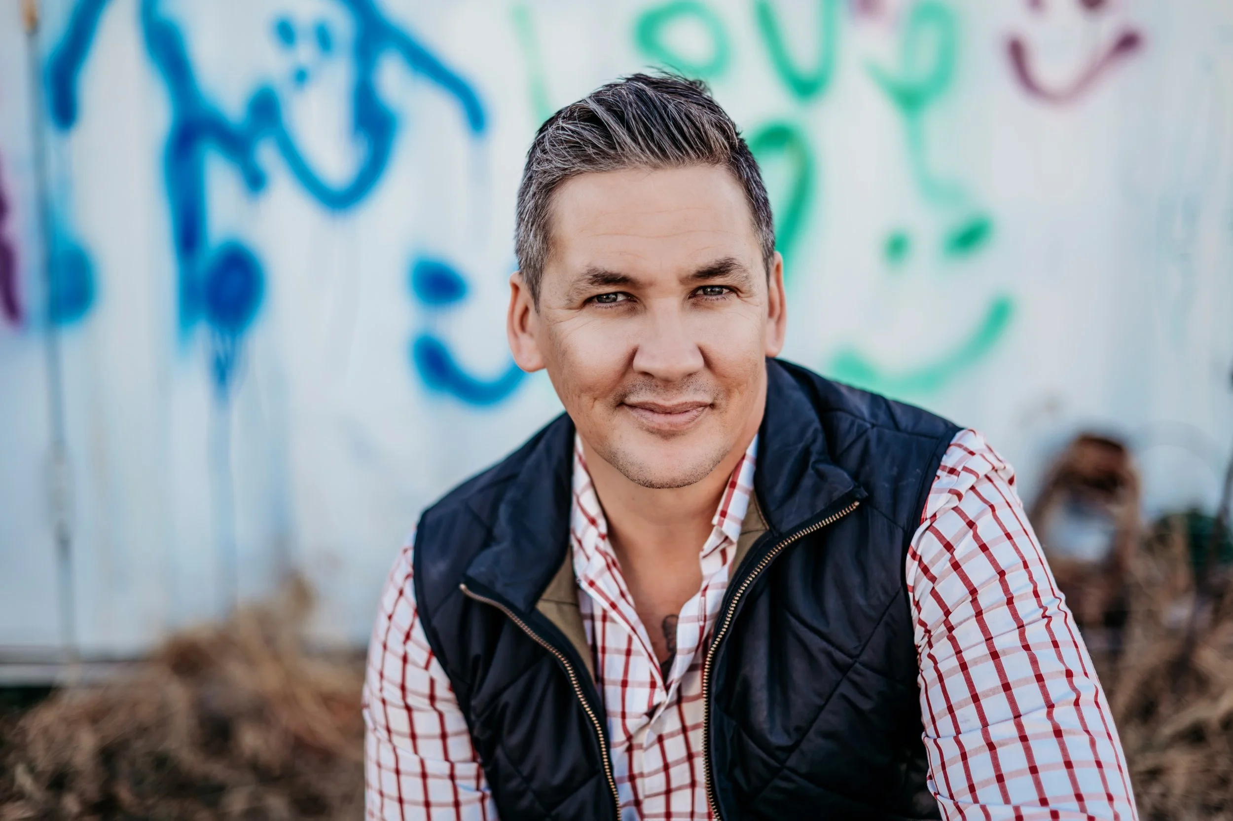 A man with gray hair and a slight smile wearing a red and white checkered shirt and a dark vest standing outdoors with a blurred background of green and blue graffiti art.