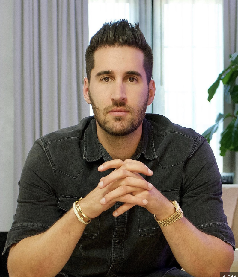 A young man with dark hair and beard sitting at a table, hands clasped, wearing a black shirt and gold wristwatch, with curtains and green plant in the background.