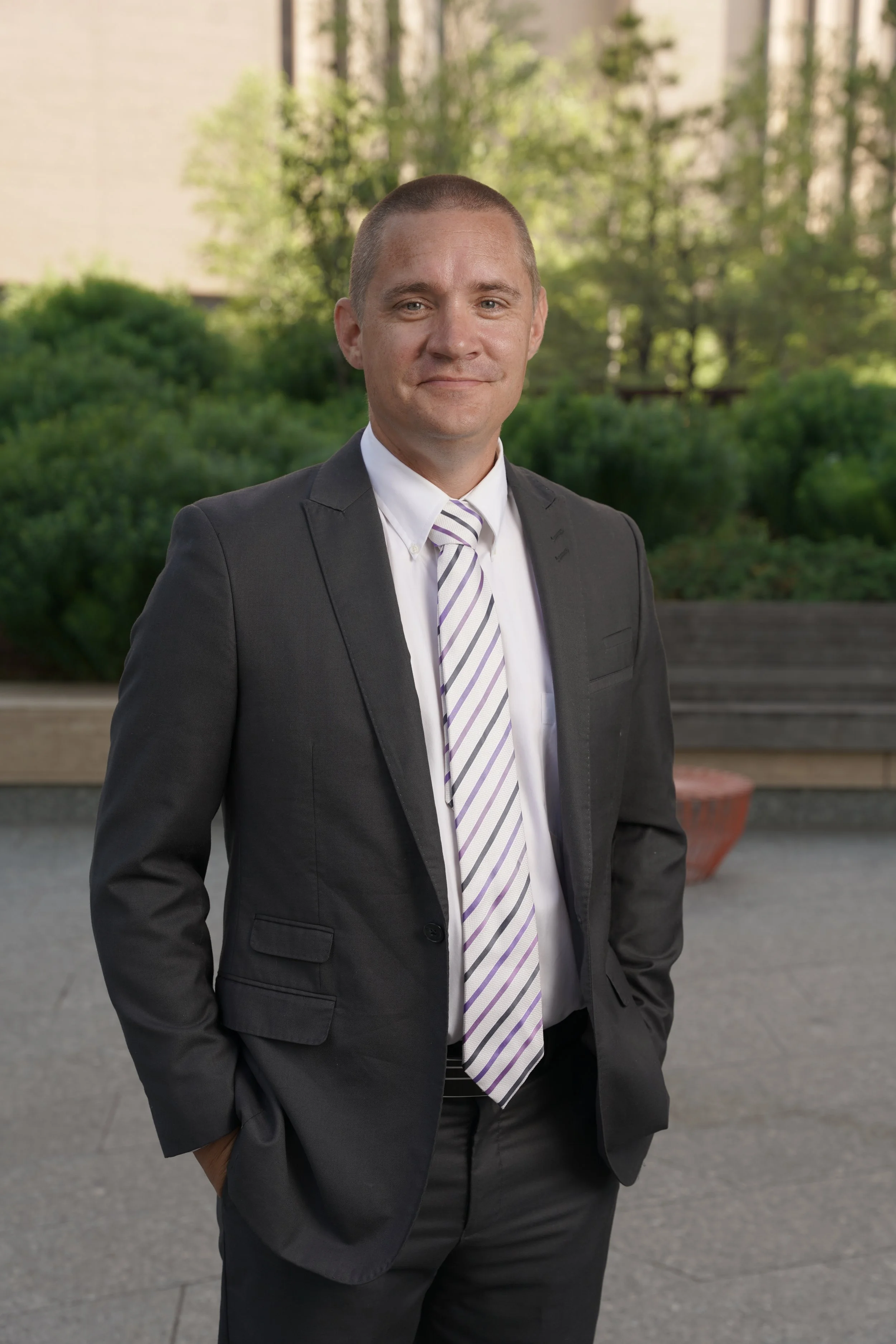 A man in a dark gray suit with a white shirt and striped purple and white tie, standing outdoors with green trees and buildings in the background.