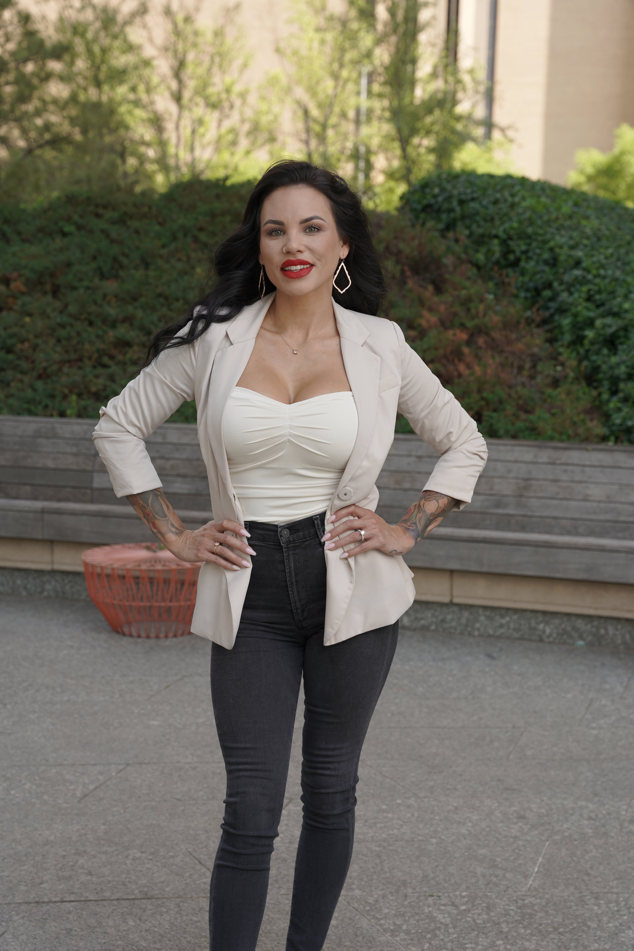 A woman with dark wavy hair, red lipstick, and tattoos on her arms, wearing a cream blazer over a white top and black jeans, posing outdoors in front of a wooden bench and green bushes.