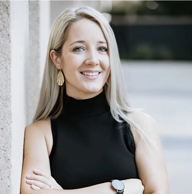 A smiling blonde woman in a black sleeveless turtleneck, wearing gold earrings and a watch, standing outdoors against a light-colored wall.