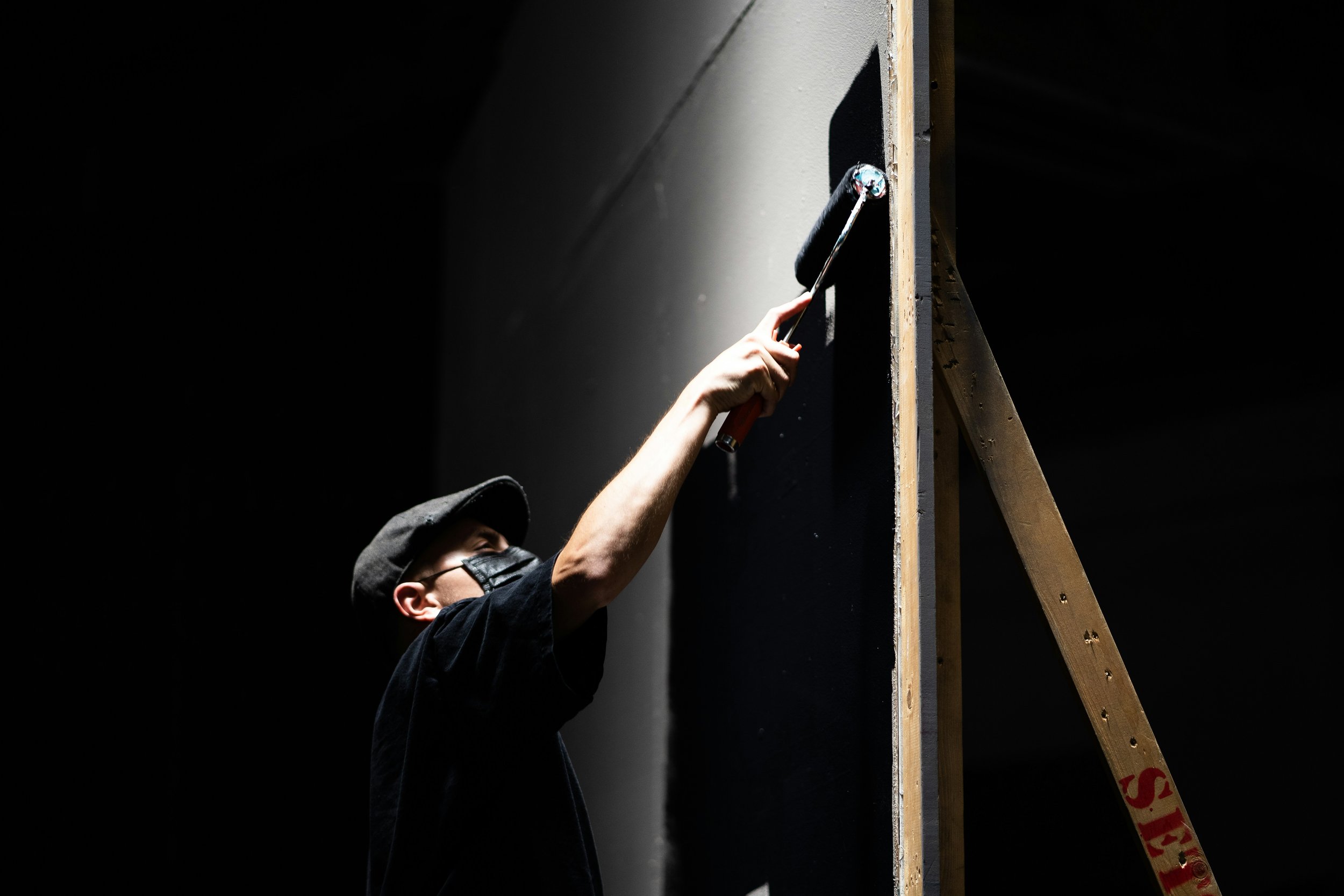 Person painting a wall with a roller brush, wearing a cap and mask, next to a wooden ladder.