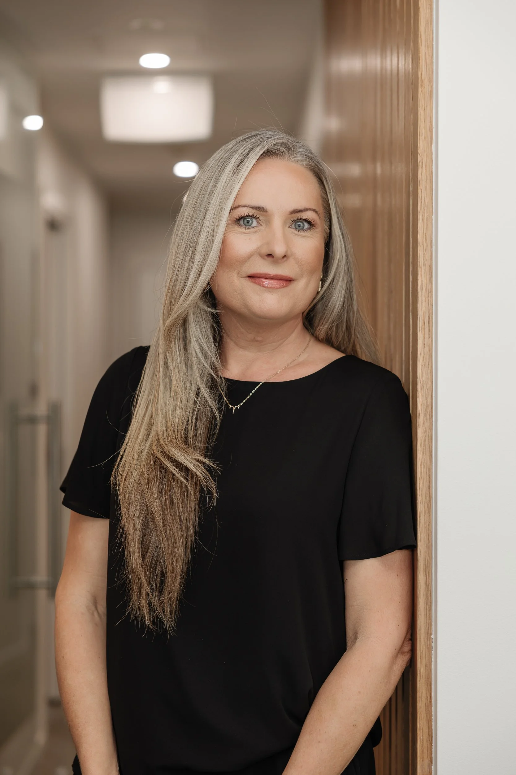 A middle-aged woman with long, silver hair and blue eyes standing in a hallway, leaning against a wooden wall and smiling gently.