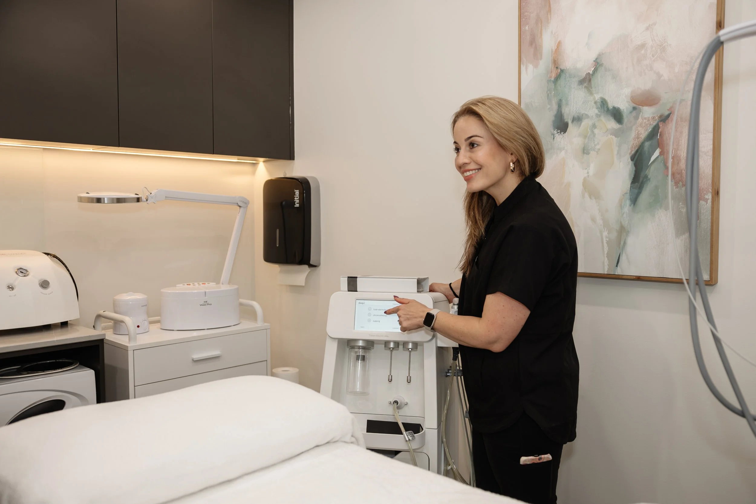 A smiling woman in black scrubs operating a medical device in a treatment room with medical equipment and an abstract painting on the wall.