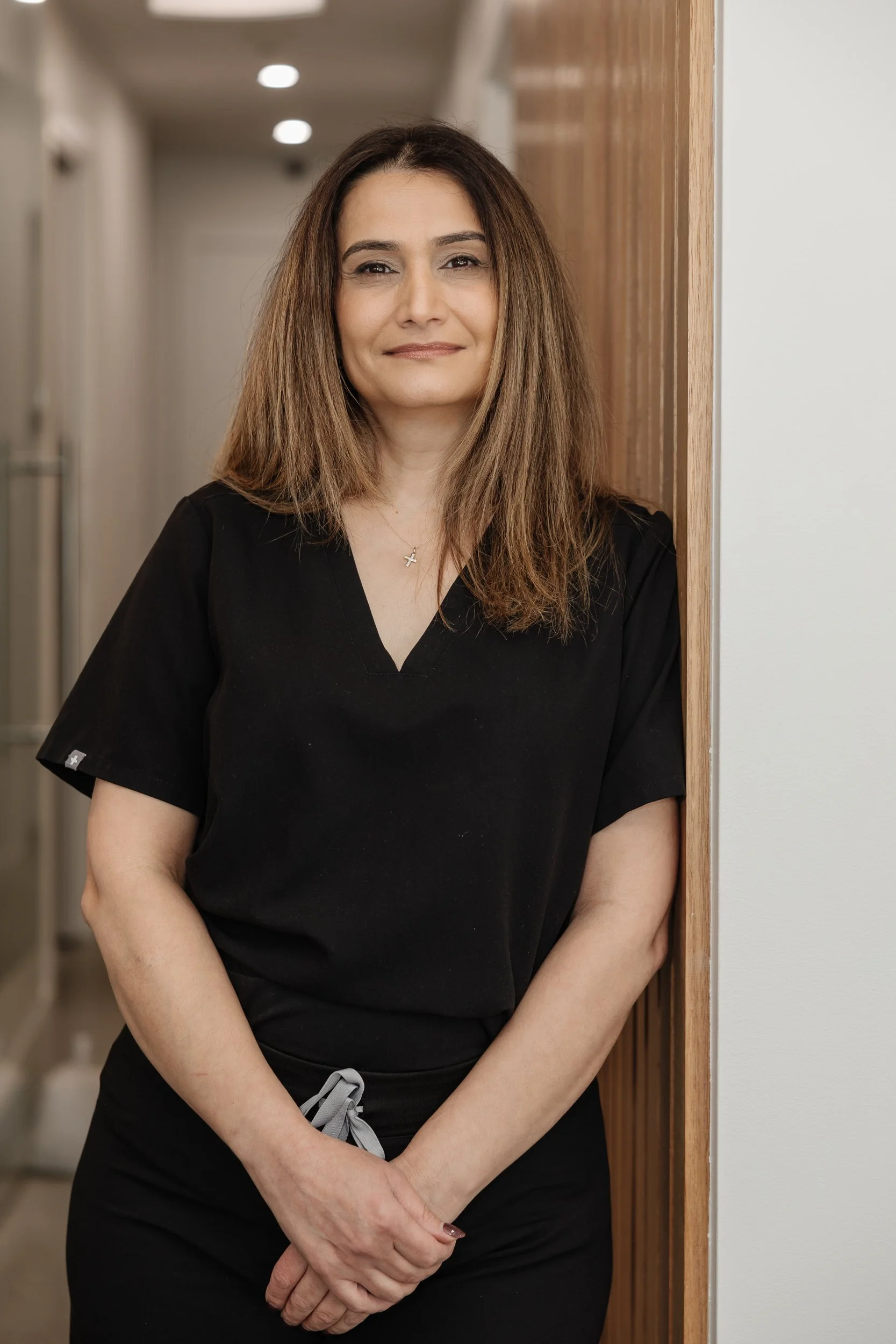A woman with long brown hair, wearing a black shirt with a small white logo on the sleeve and a necklace, standing indoors next to a wooden wall.