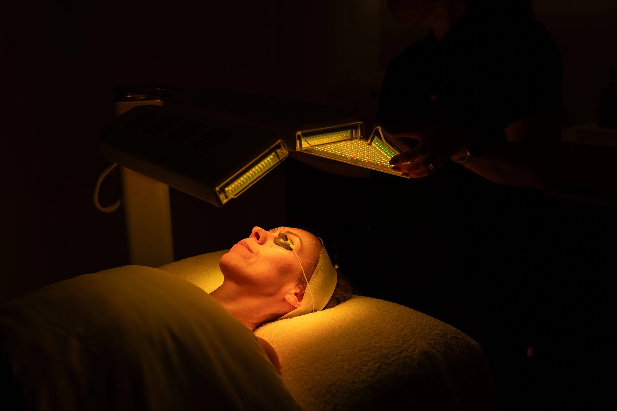 A person undergoing a medical or cosmetic procedure, lying down with a protective eye covering, illuminated by a specialized treatment device in a dark room.