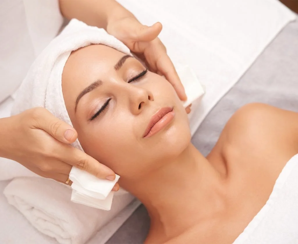 A woman receiving a facial massage in a spa, with her eyes closed and a white headband on.