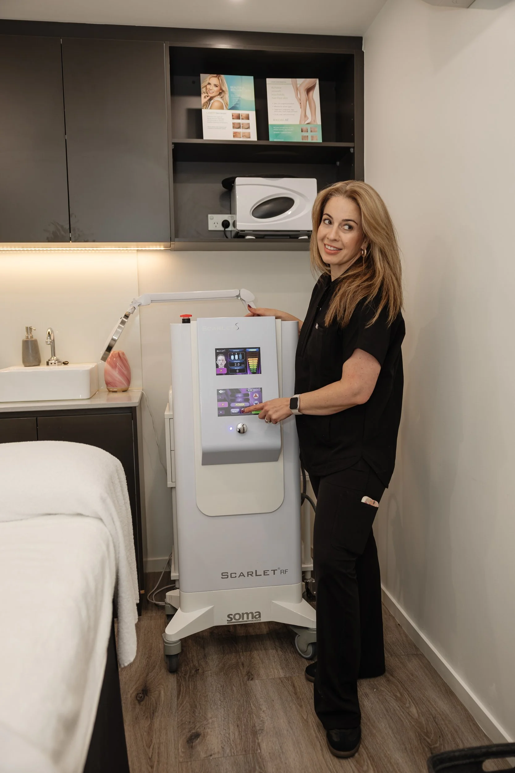 A woman in black scrubs standing next to a medical device labeled 'Scarlet RF' in a room with white walls and a wooden floor, with part of a treatment bed visible in the foreground.