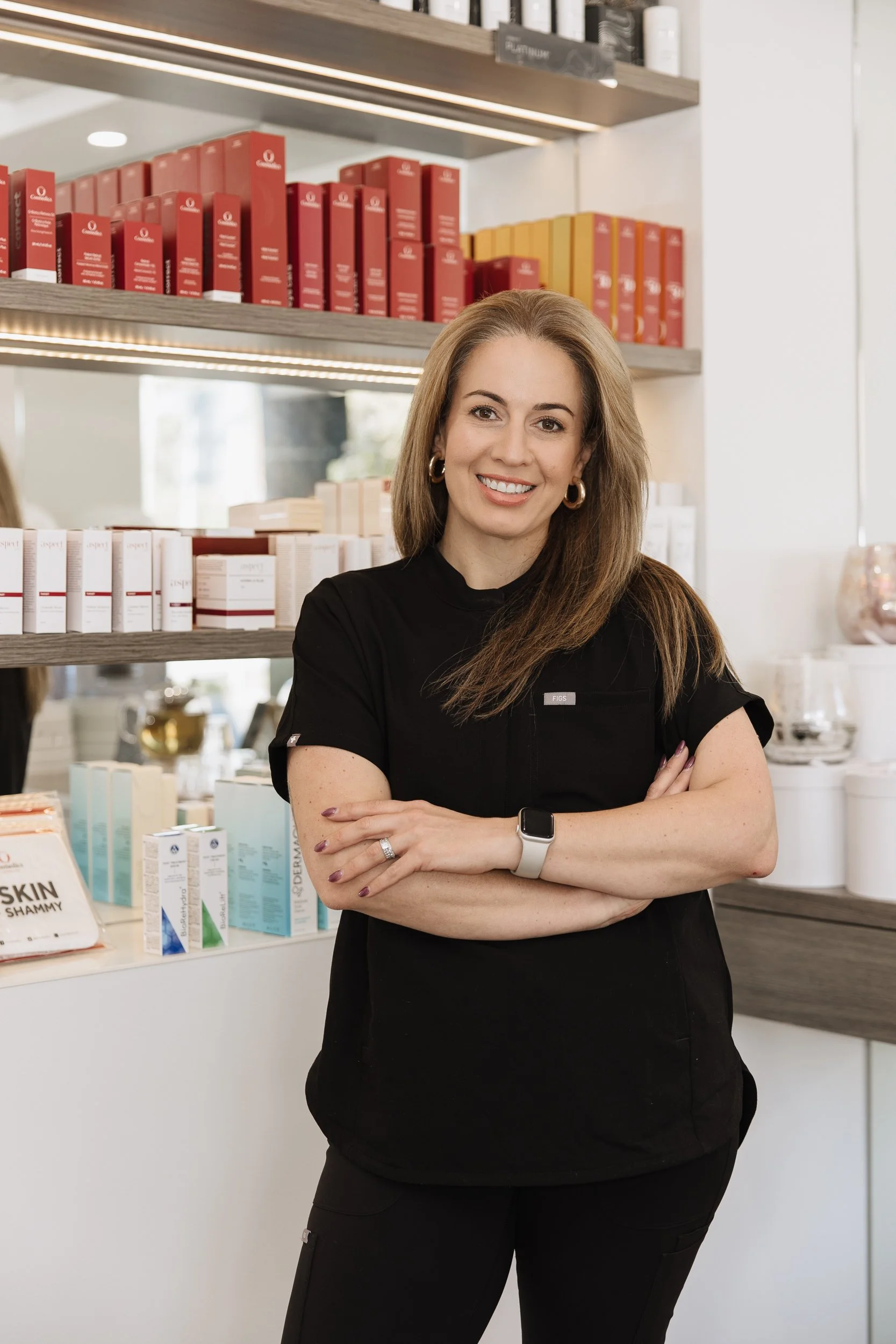 A smiling woman with light brown hair wearing a black shirt and black pants, standing with her arms crossed in front of shelves filled with skincare products in a retail store.
