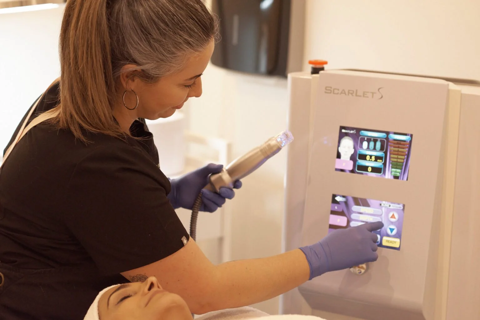A woman with gray hair, wearing purple gloves, is operating a medical device with a touchscreen interface, next to a patient lying down with eyes closed.