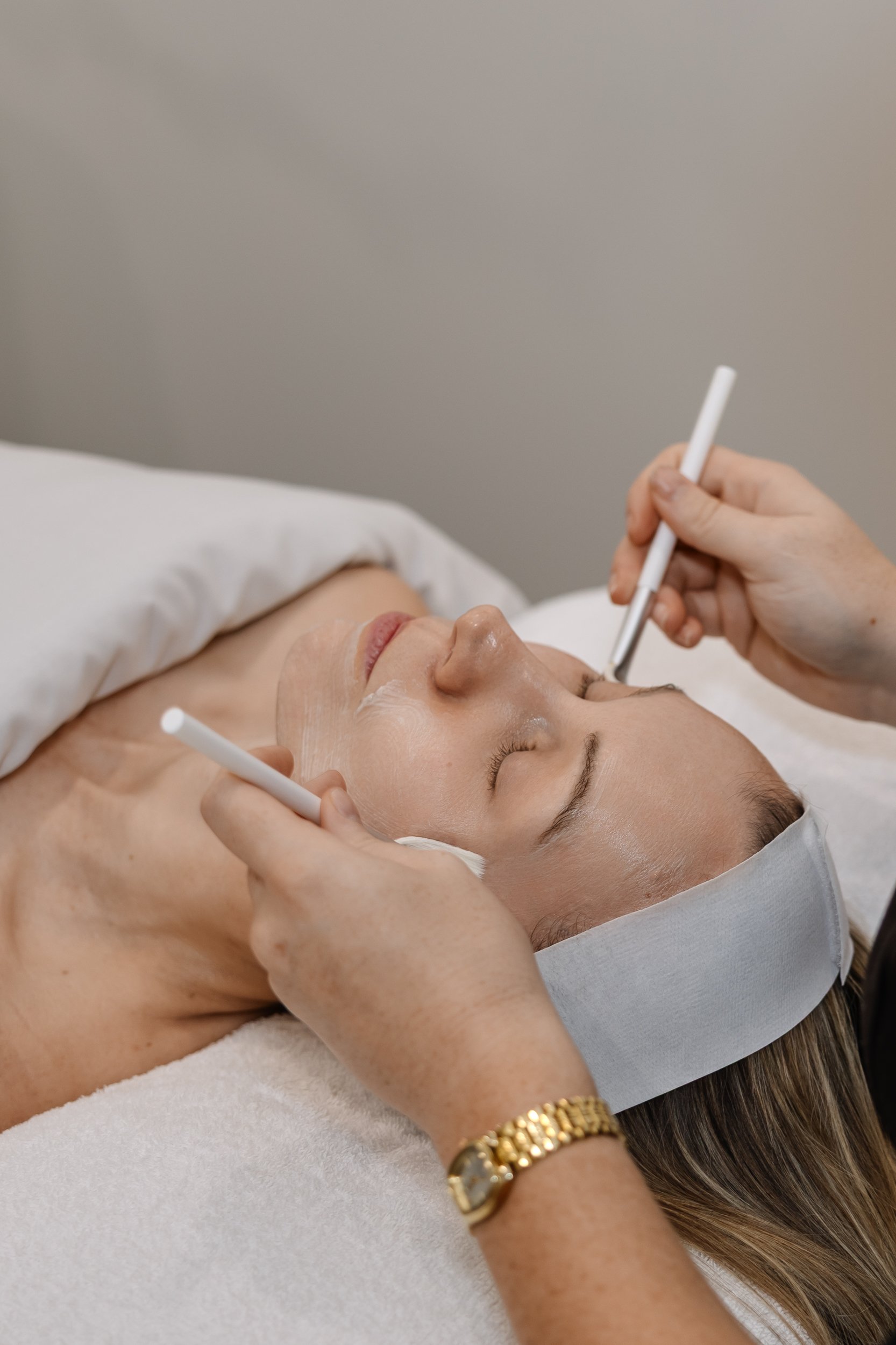 A woman lies with eyes closed on a treatment bed while a skincare professional applies a facial mask and uses a tool on her face in a spa or medical setting.