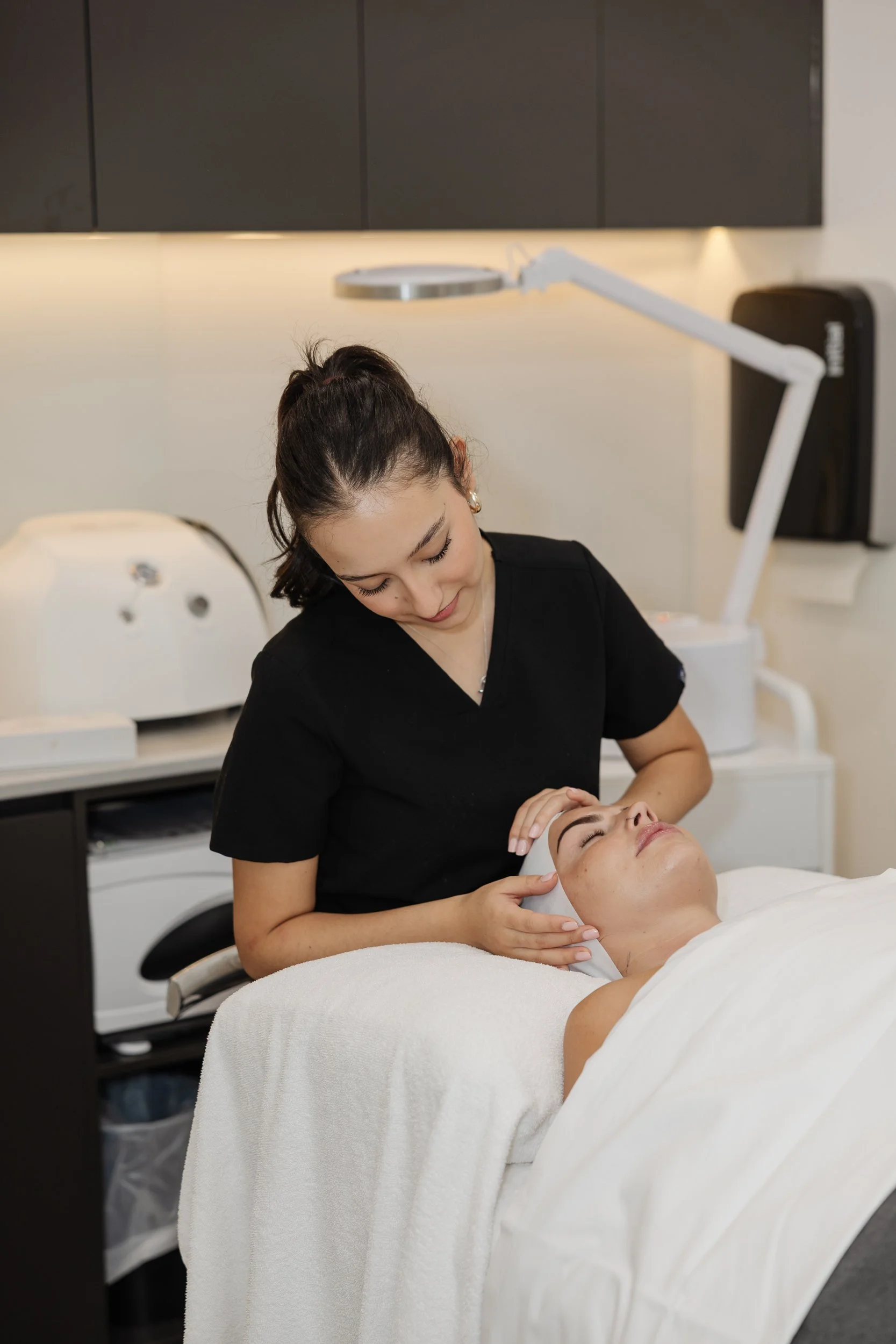 A woman receiving a facial treatment from a skincare specialist in a spa or clinic setting, with skincare equipment visible in the background.