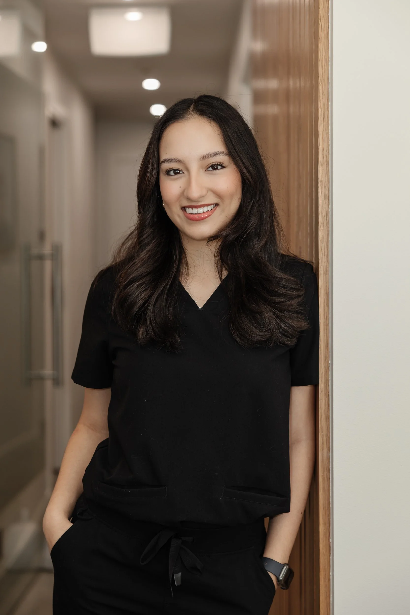 A young woman with long dark hair, wearing a black top, standing with her hand in her pants pocket, smiling and leaning against a wooden wall in an indoor hallway with ceiling lights.