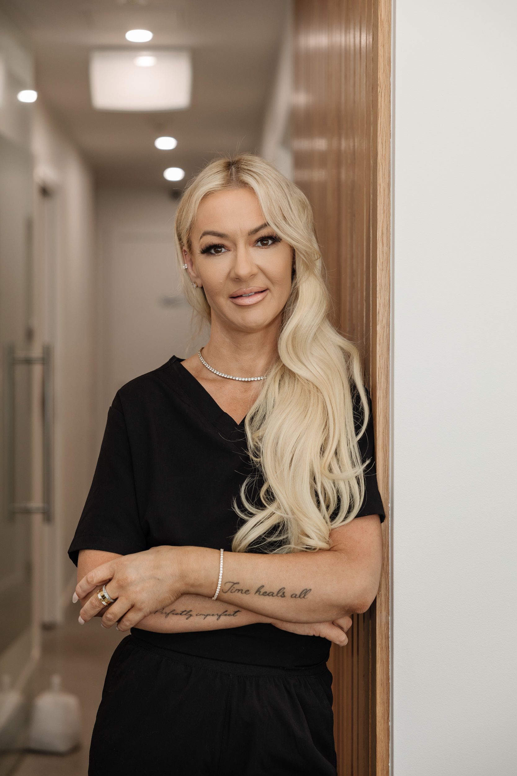 A blonde woman with long, wavy hair standing in a hallway, wearing a black top, jewelry, and with tattoos on her forearm.