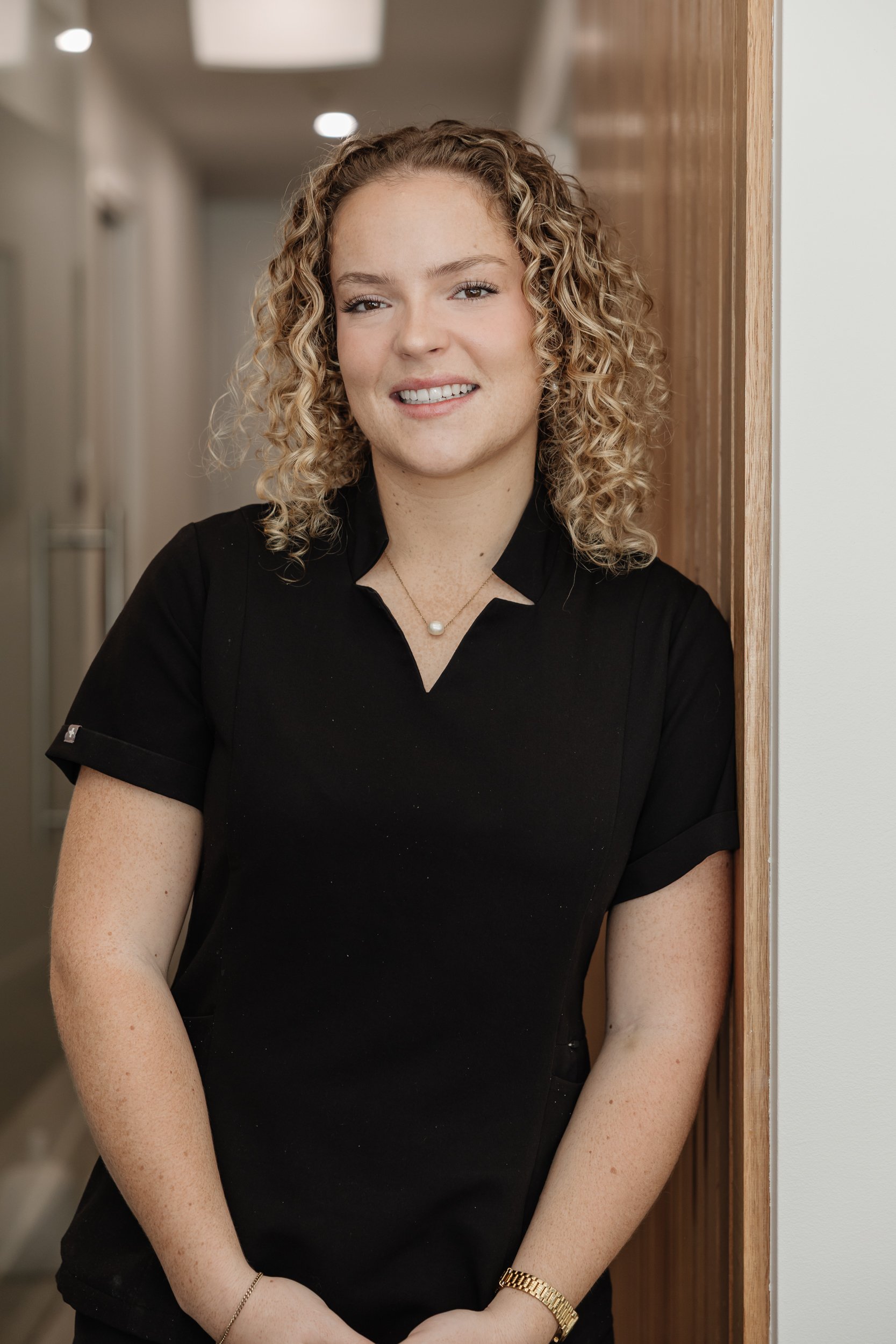 A woman with curly blonde hair smiling and leaning against a doorframe in a professional setting, wearing a black top, a pearl necklace, a gold watch, and a bracelet.