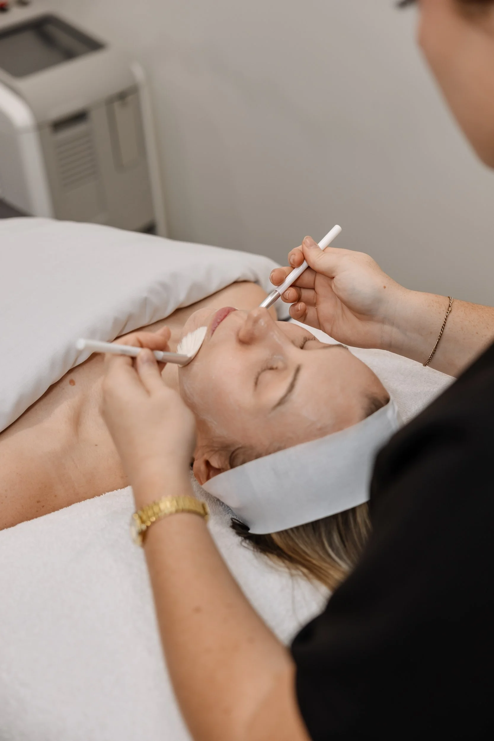 A woman is lying on a bed receiving a facial treatment, with a practitioner applying a product to her face using a tool.