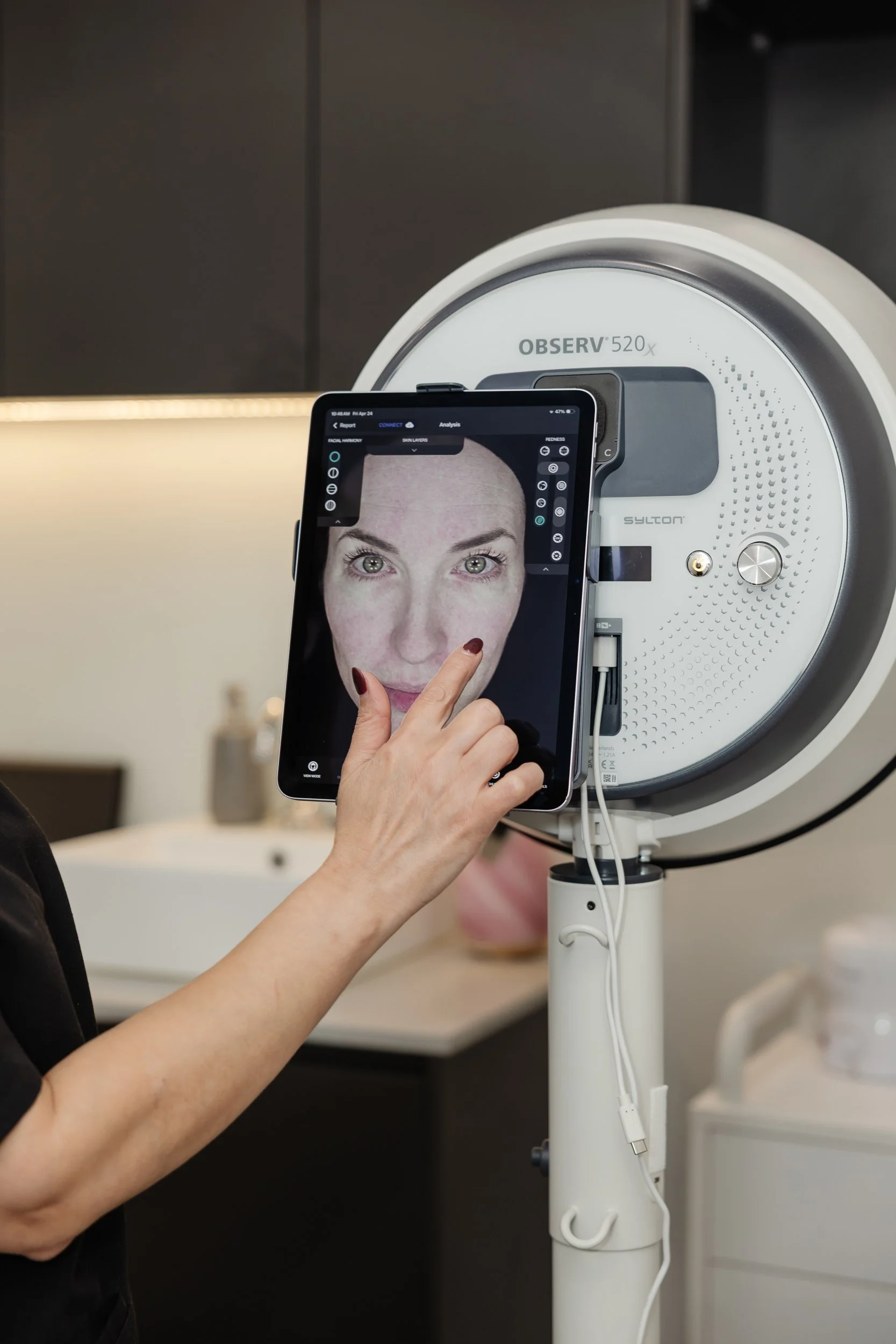 A woman with painted nails points to her face on a smartphone screen, which is aligned with a medical imaging device in a clinical setting.