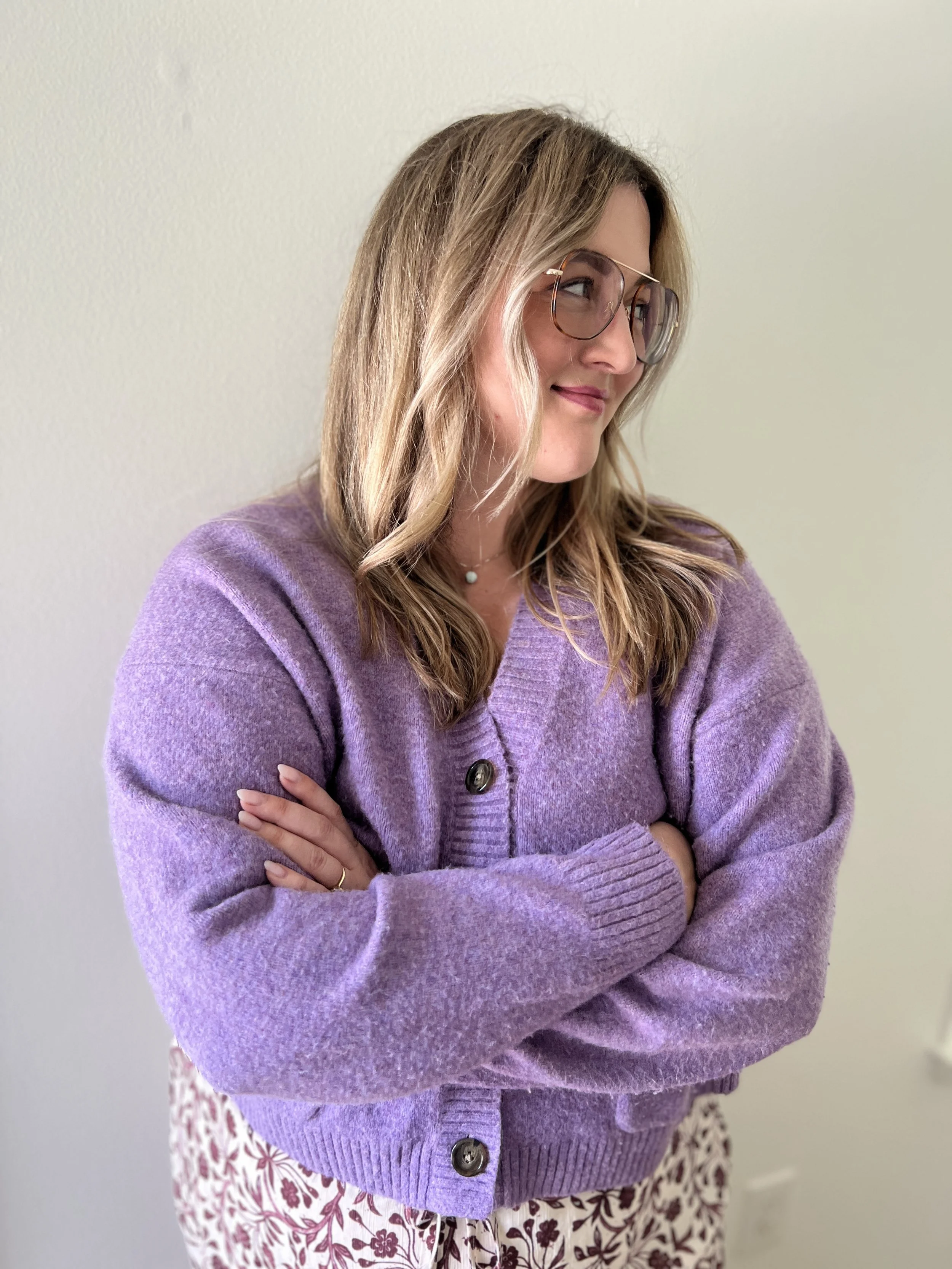 A woman with blonde hair wearing glasses, a purple cardigan, and floral-patterned pants, standing with arms crossed against a plain white wall.