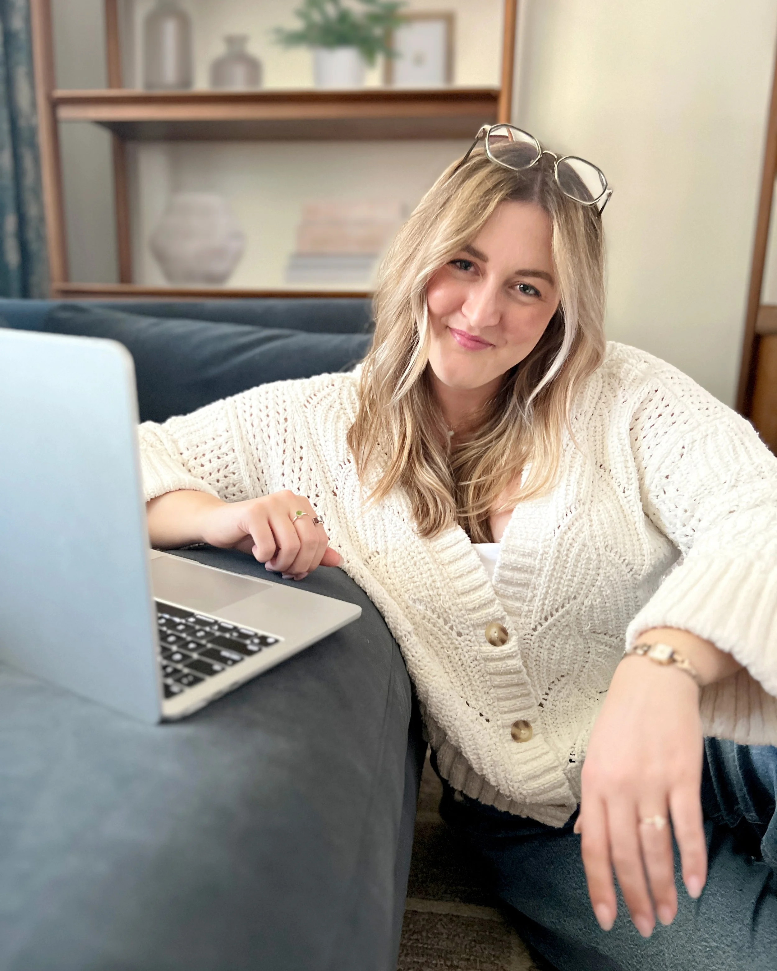 A woman with blonde hair and glasses resting on her head, wearing a knitted cream-colored sweater, is sitting at a table with a laptop. She is smiling and looking at the camera, with her right hand resting on the table and wearing a ring and a bracelet. The background features a blurred bookshelf with decorative items and books.