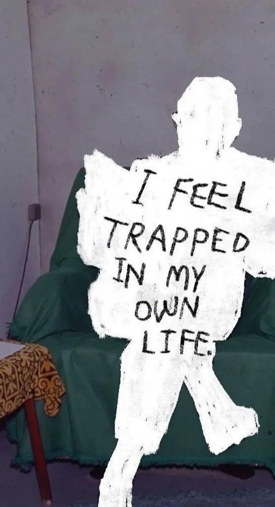 A person sitting on a green chair with a marker-drawn white silhouette overlay and the words "I FEEL TRAPPED IN MY OWN LIFE" written on it.