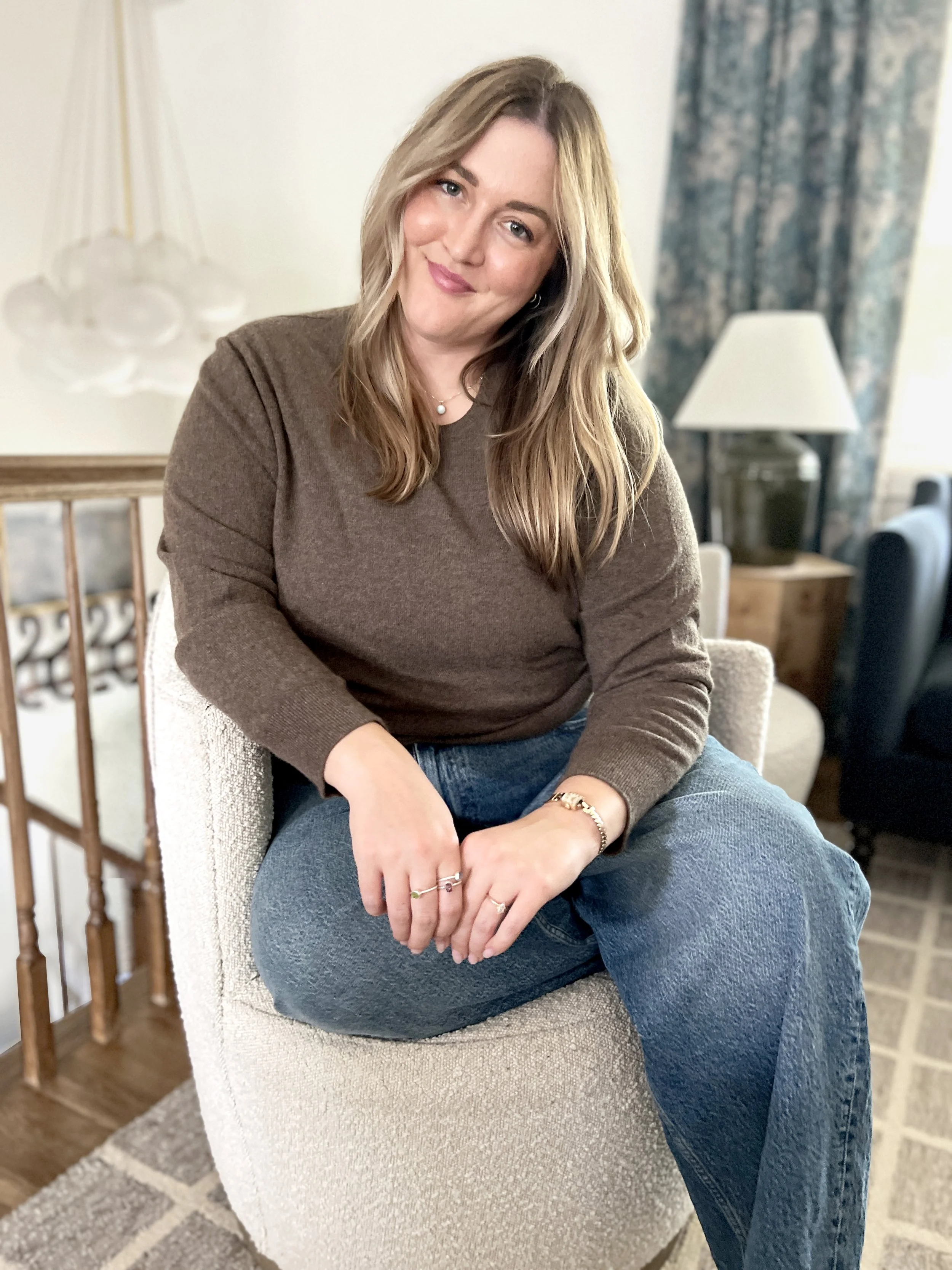 A woman with long blonde hair sitting on a beige armchair in a cozy living room, smiling at the camera, wearing a brown sweater and blue jeans, with a background featuring a table lamp, curtains, and a staircase railing.