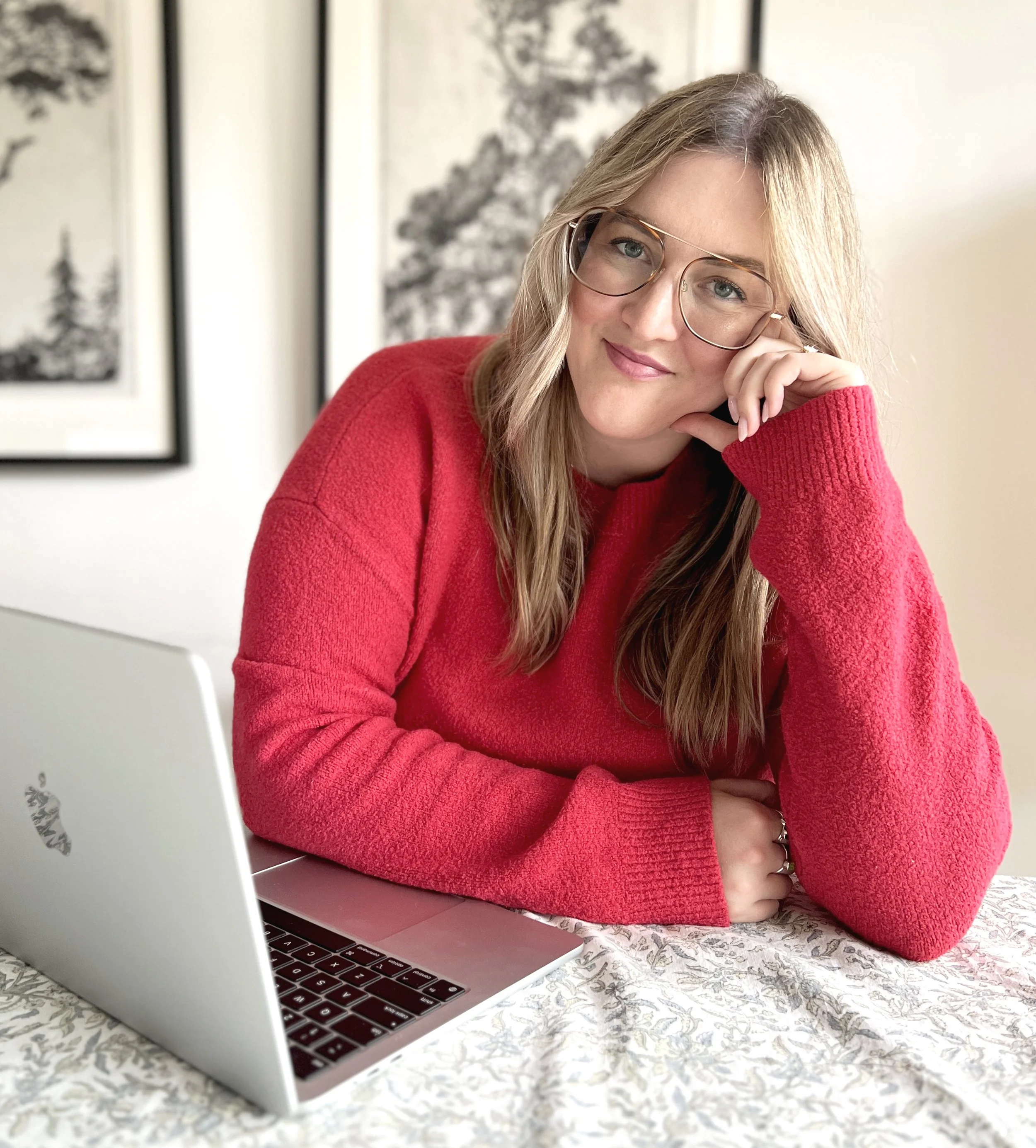 A young woman with blond hair and glasses wearing a red sweater is sitting at a table with a laptop, resting her face on her hand and looking at the camera with a slight smile in a room with framed artwork in the background.