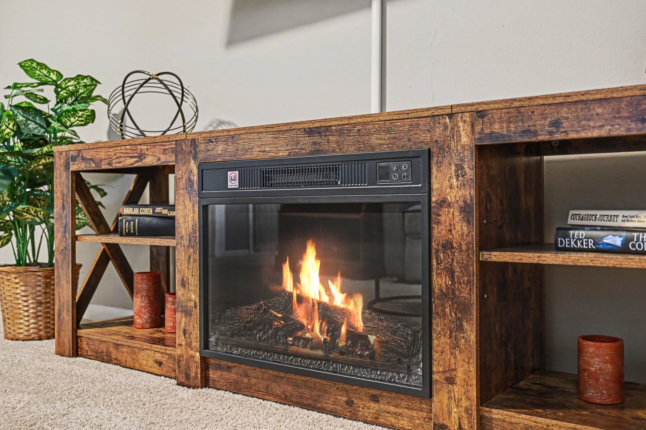 An electric fireplace mantel with a virtual fire burning inside the fireplace, with a silver geometric sculpture and a potted plant, and books in a shelf underneath, located in a cozy living room.