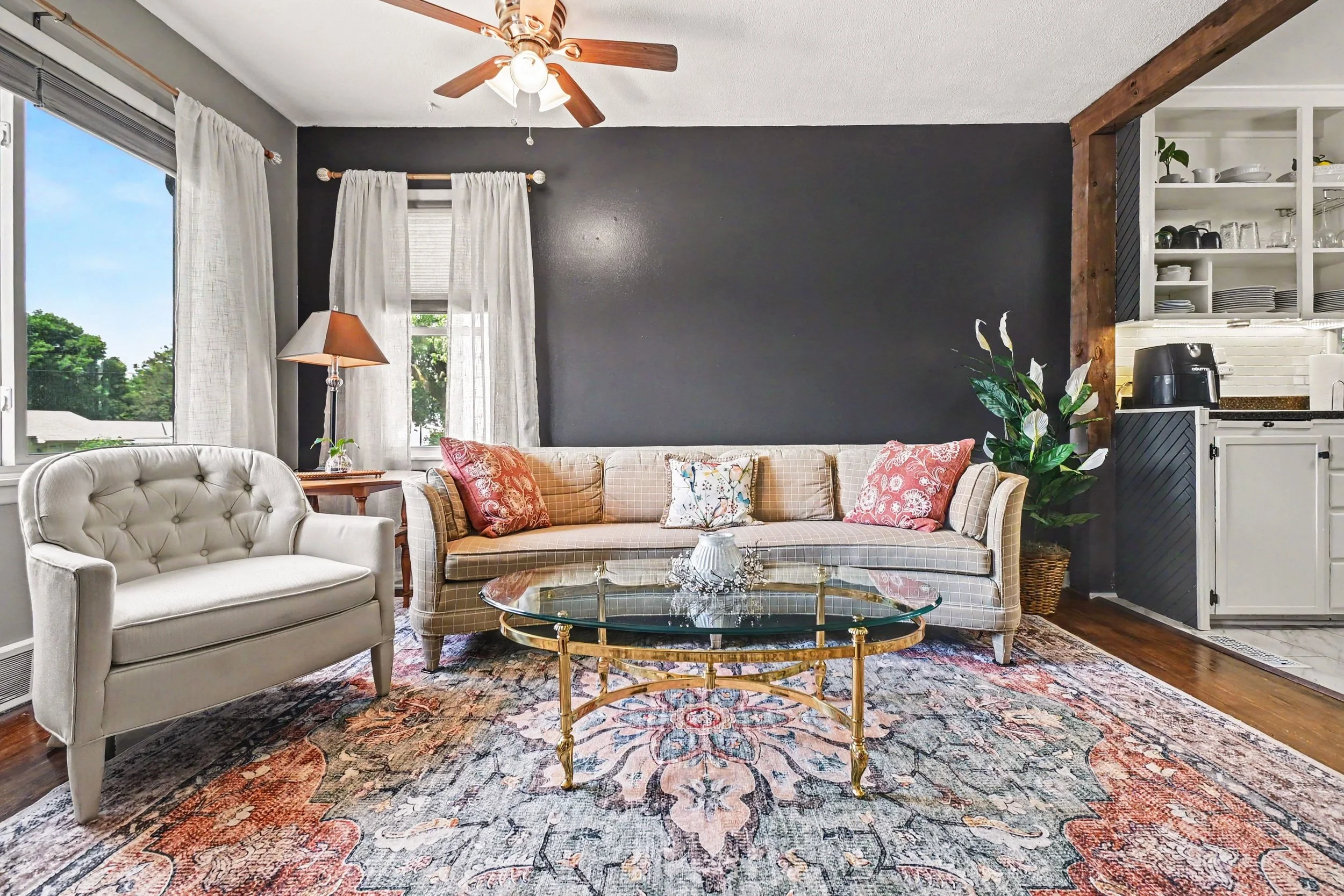 Living room with beige and floral-patterned couch, glass coffee table, white armchair, colorful area rug, dark accent wall, and large windows with white curtains. A ceiling fan with lights is mounted on the ceiling. Kitchen area partially visible on 