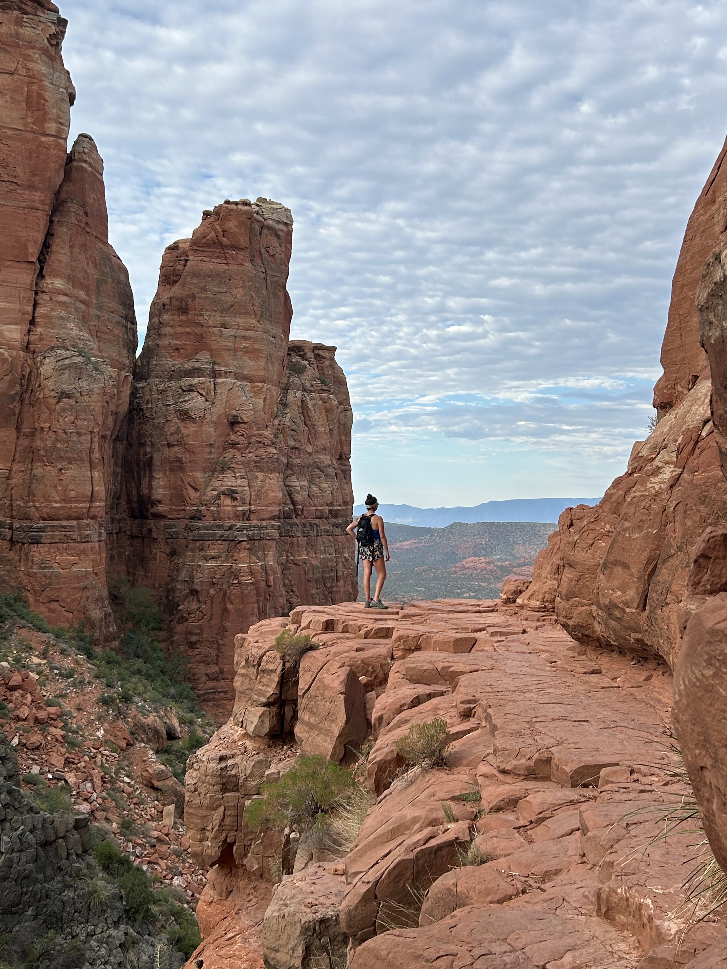 A person standing on a rocky ledge among large red rock formations in a canyon, with a view of distant mountains and a cloudy sky overhead.