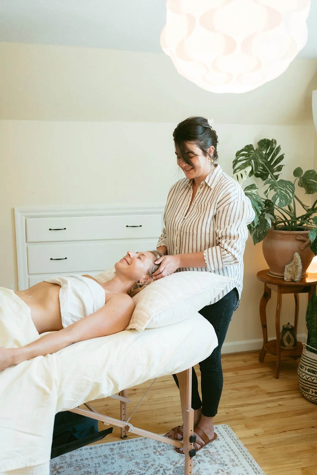 A woman receiving a head massage from a massage therapist in a cozy, well-lit room with plants and wooden furniture.