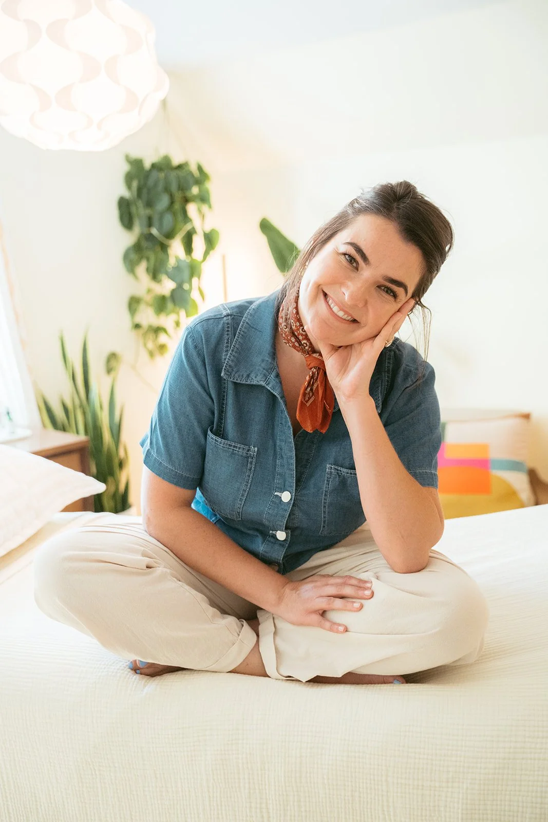 A woman with dark hair and a friendly smile, sitting cross-legged on a bed in a sunny, cozy bedroom, wearing a denim shirt and a neck scarf.