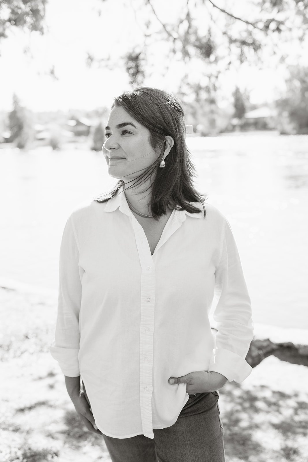 A woman with shoulder-length dark hair, wearing a white button-up shirt and earrings, standing outdoors with a background of trees and snow.