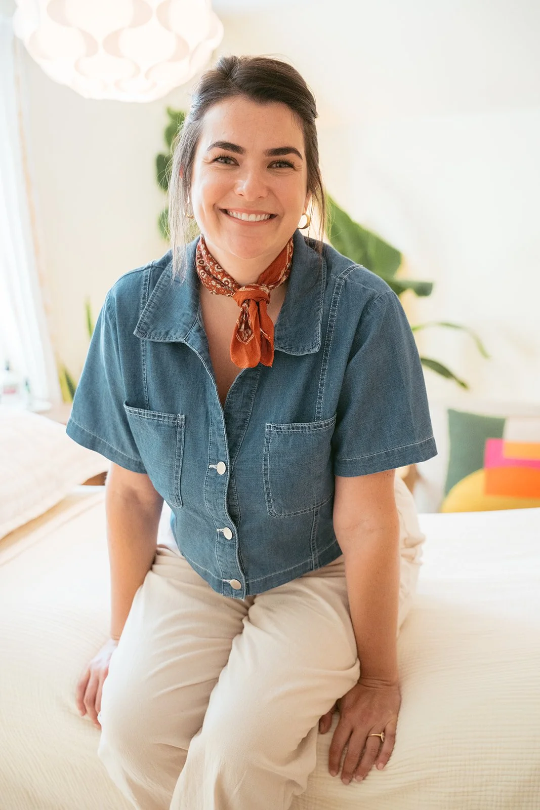 A smiling woman with dark hair, wearing a denim shirt and a red patterned neck scarf, sitting on a bed with colorful pillows in a bright room with plants and a modern ceiling light.
