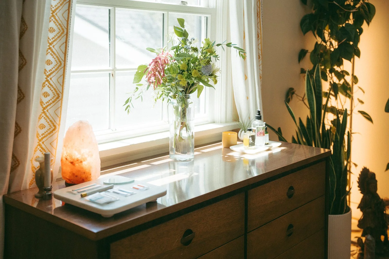 Decorative wooden dresser with a salt lamp, a tray of skincare products, a glass vase with fresh flowers, and various small containers near a window with sunlight and curtains, surrounded by indoor plants.