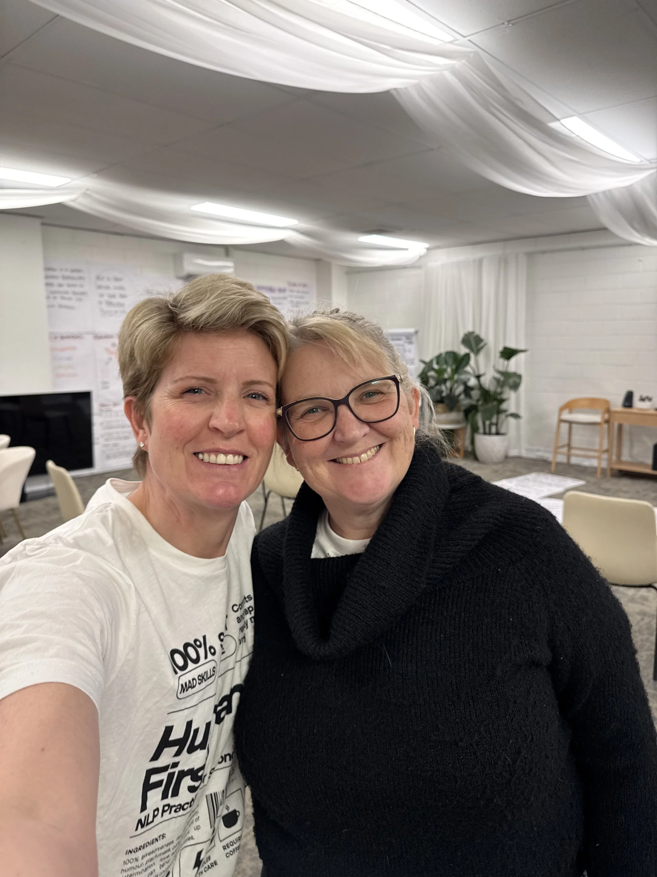Two women smiling and taking a selfie in an indoor room with white ceiling decorations, potted plants, and chairs in the background.