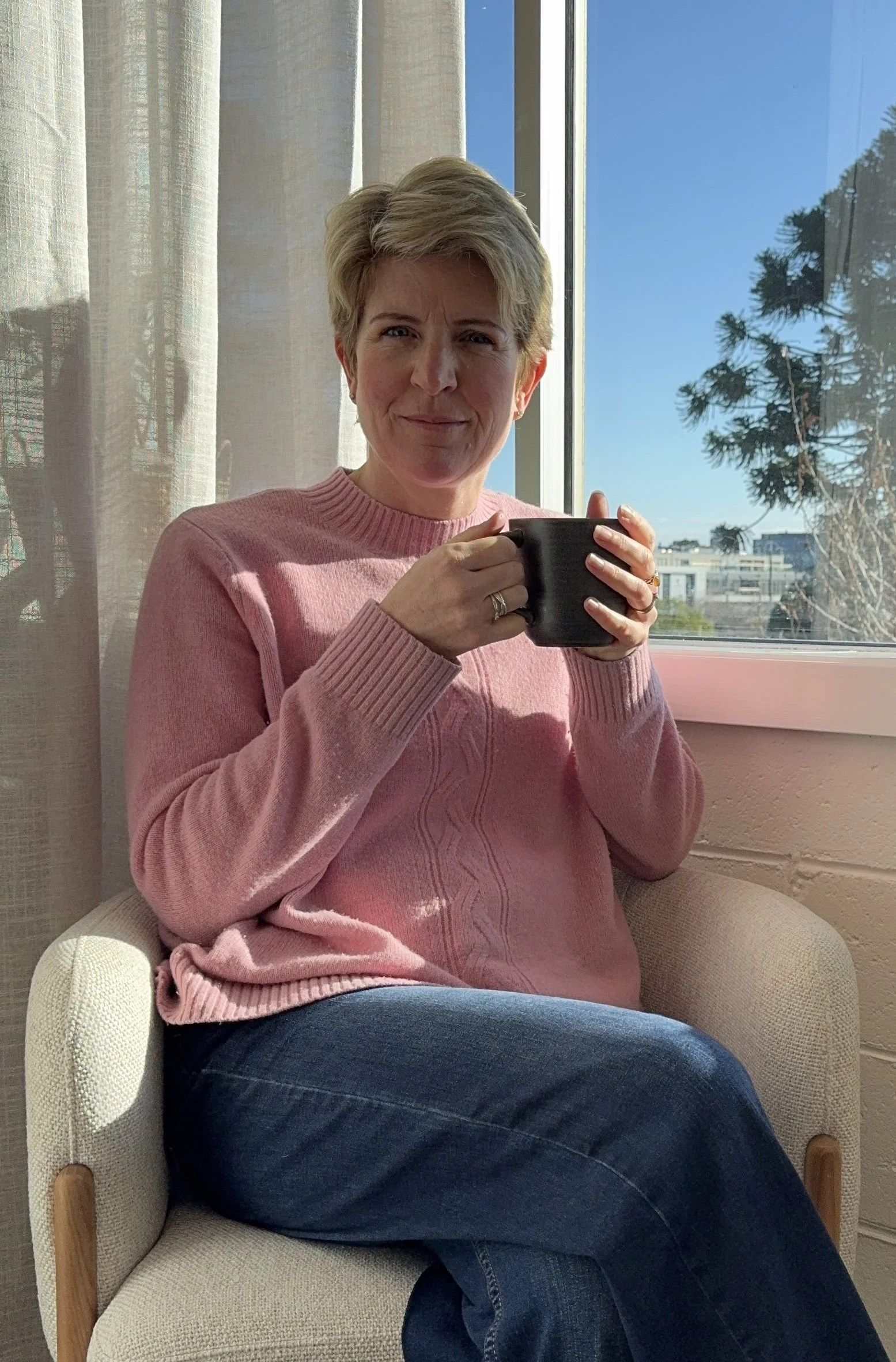 A woman with short blonde hair, wearing a pink sweater, sitting on a beige armchair near a window with white curtains. She is holding a black mug and smiling at the camera. Outside the window, there are trees and a clear blue sky.