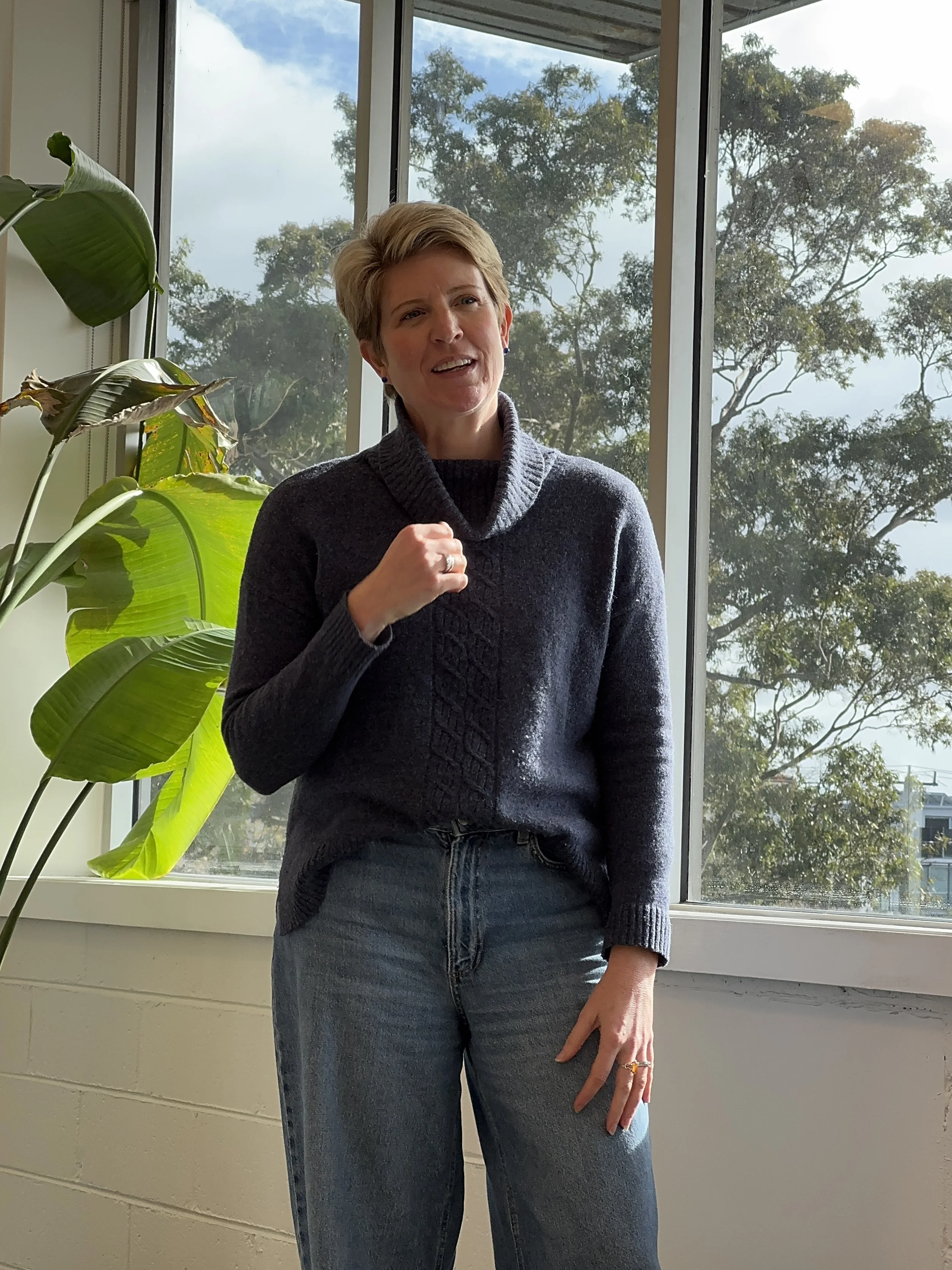 A woman with short blonde hair wearing a dark gray sweater and blue jeans standing near a large window with outdoor trees visible behind her.