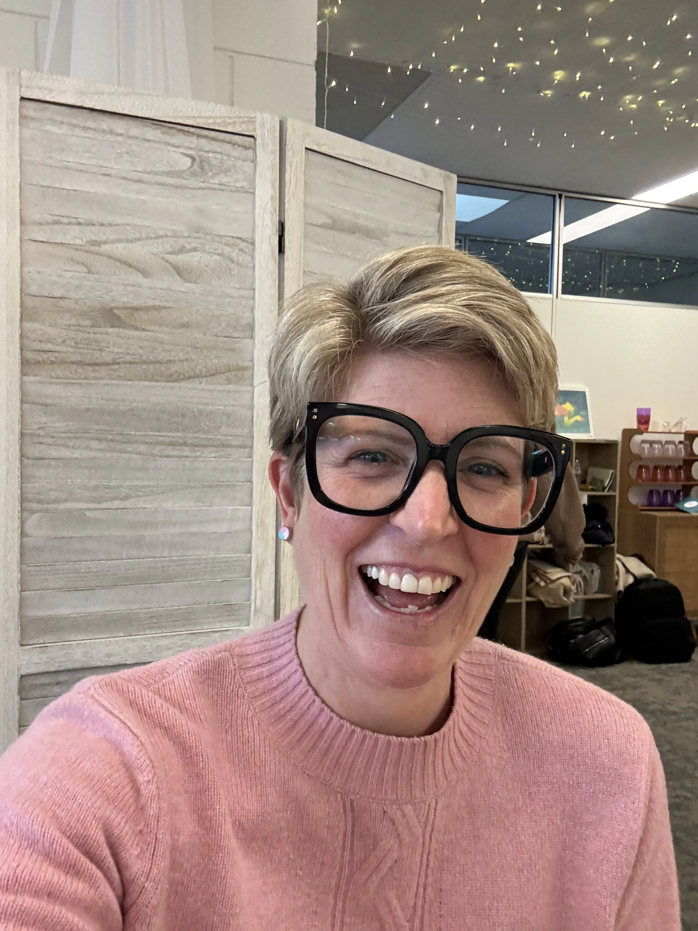 A smiling woman with short blonde hair and black glasses wearing a pink sweater. In the background, there are shelves with various items, a small colorful painting, and a room with fairy lights on the ceiling, partially visible through a glass window.