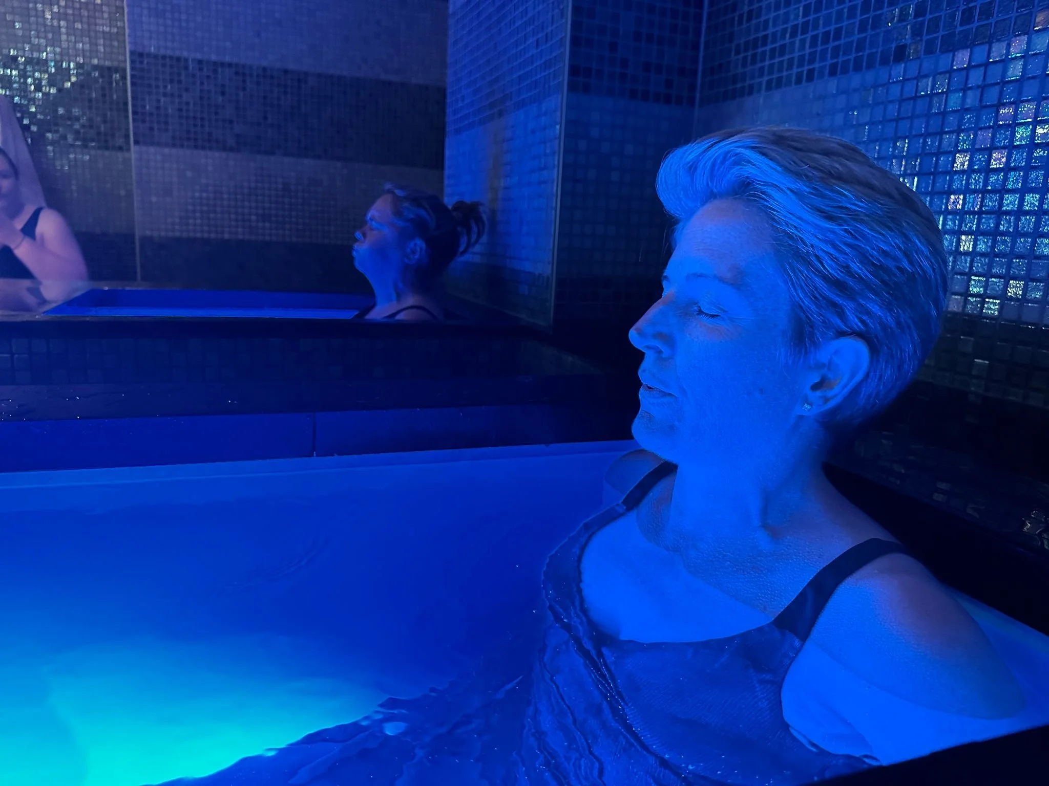 Women relax in a blue-lit spa pool with tiled walls.