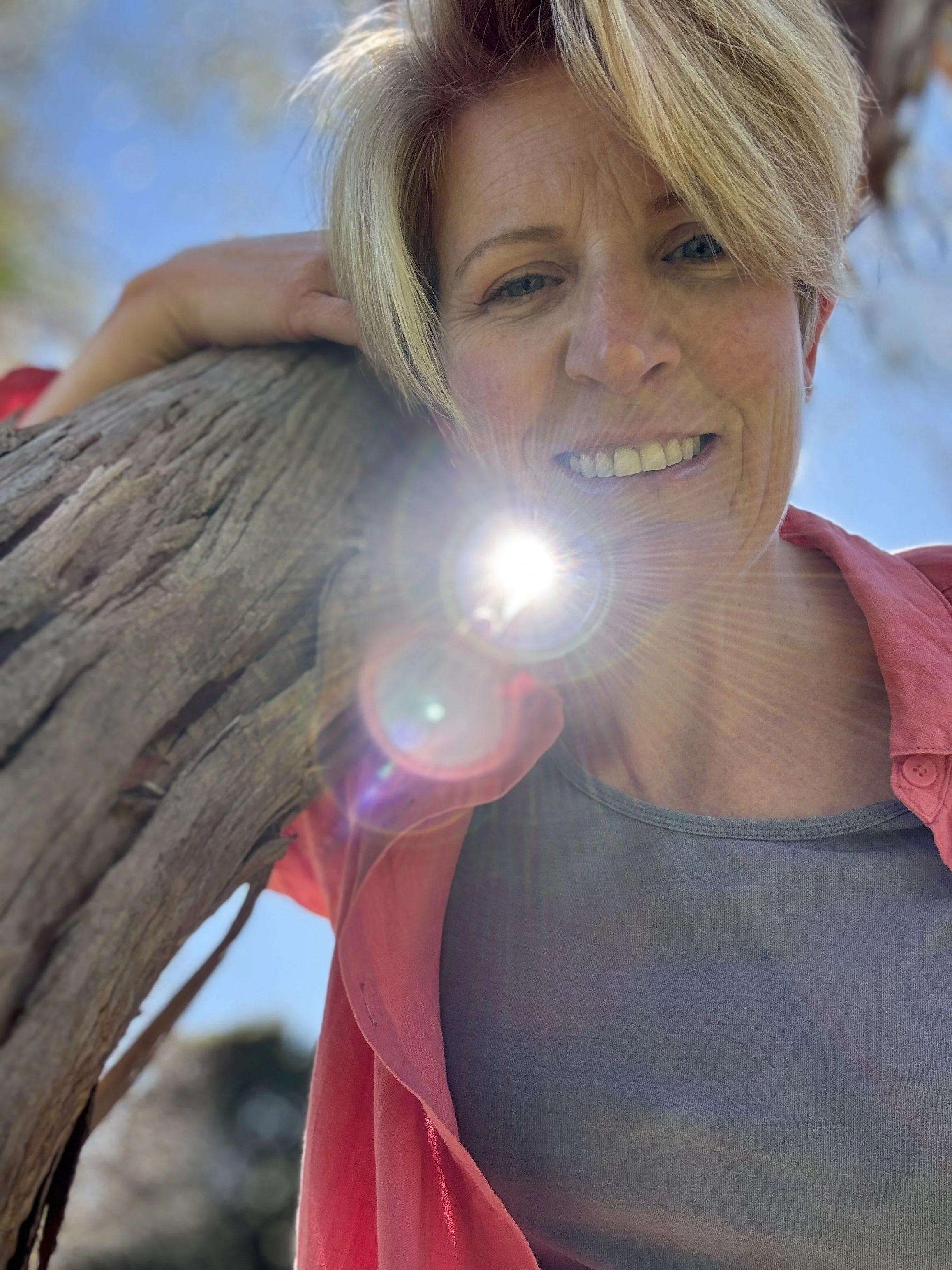 A woman with blonde hair leaning on a tree trunk with the sun shining on her face, blue sky in the background.