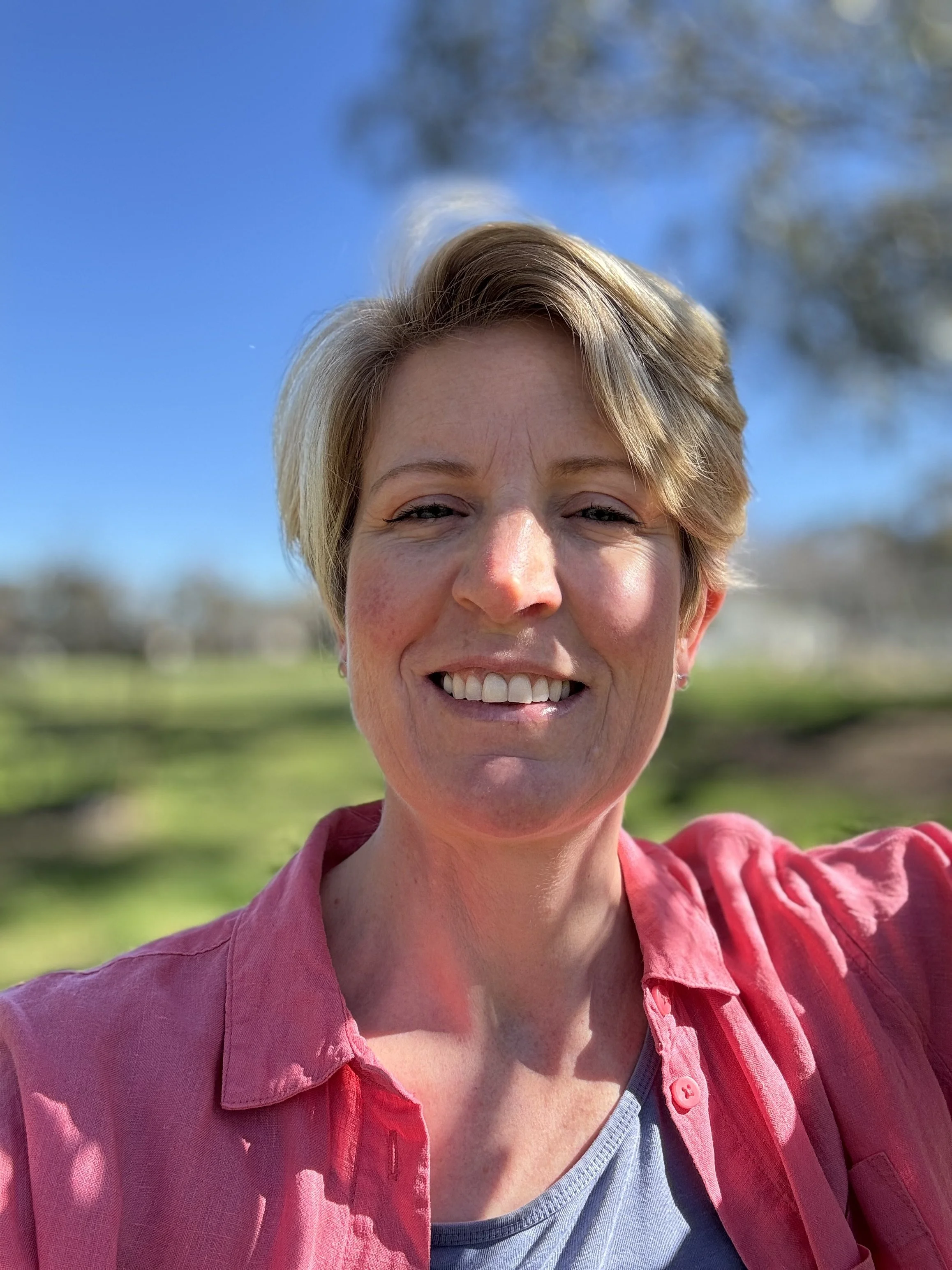 A smiling woman with short blonde hair wearing a pink shirt and gray top, outdoors with a clear blue sky and green landscape in the background.