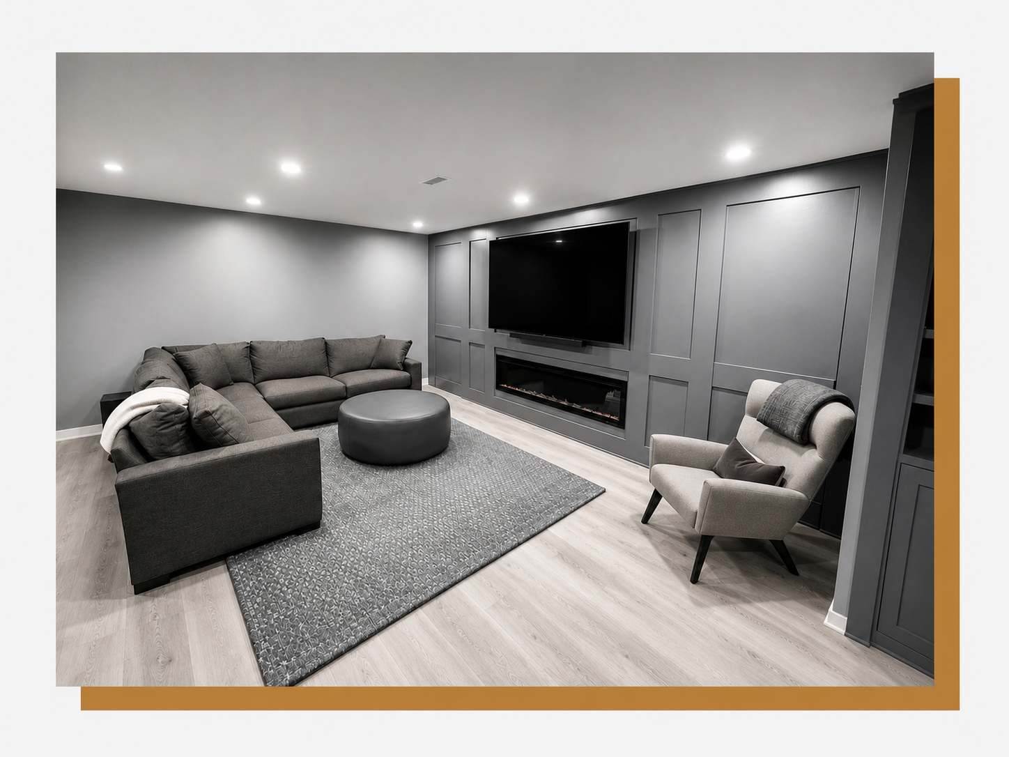 Modern living room with gray walls, gray sectional sofa, gray armchair, black ottoman, large flat-screen TV, electric fireplace, gray rug, ceiling lights, hardwood floors.