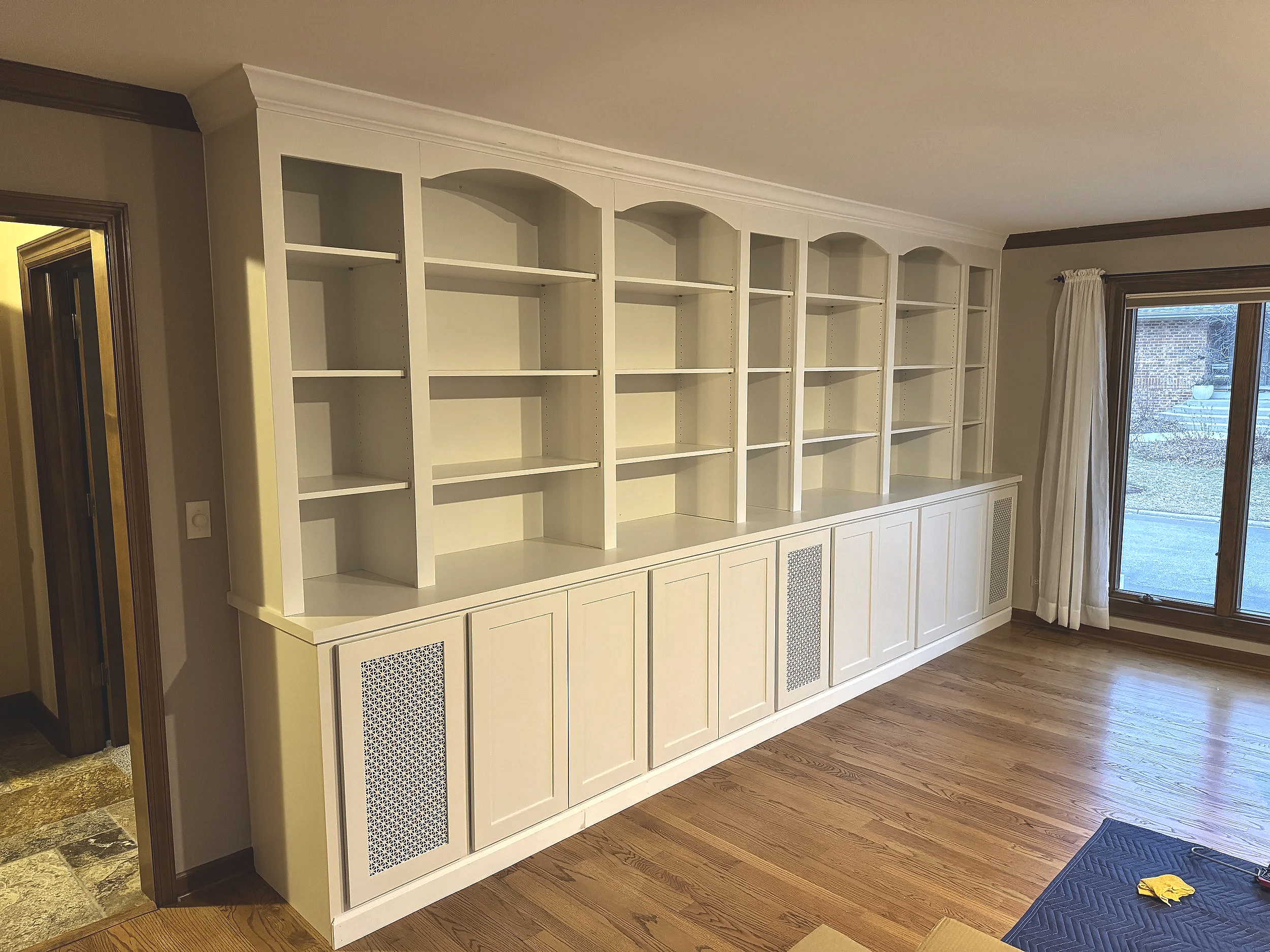 Empty white built-in bookshelf with shelves and cabinets below, located in a room with hardwood floors and a large window with white curtains.