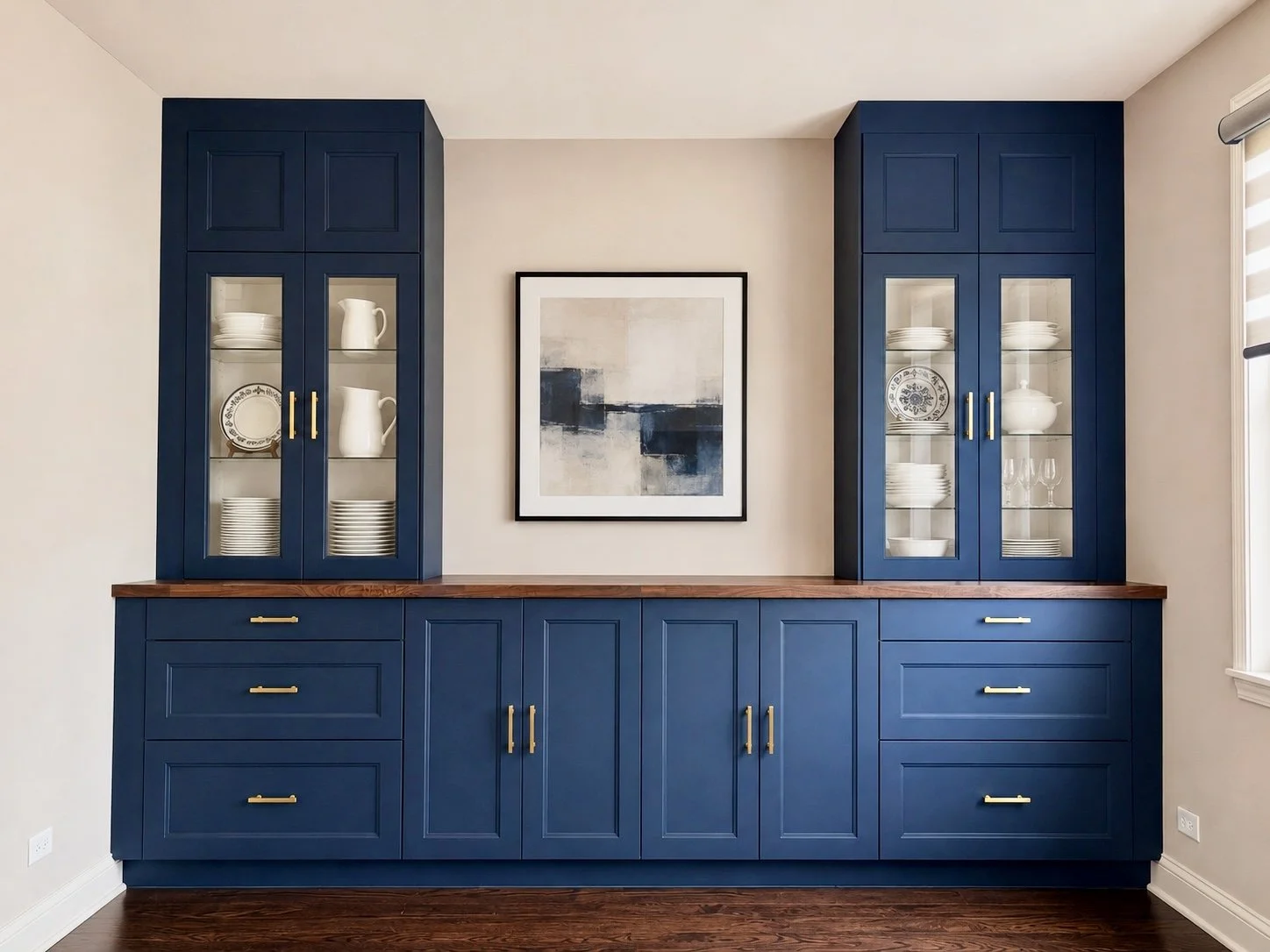 Blue built-in kitchen cabinets with glass doors, gold handles, and a wooden countertop, with a framed abstract art piece on the wall above.