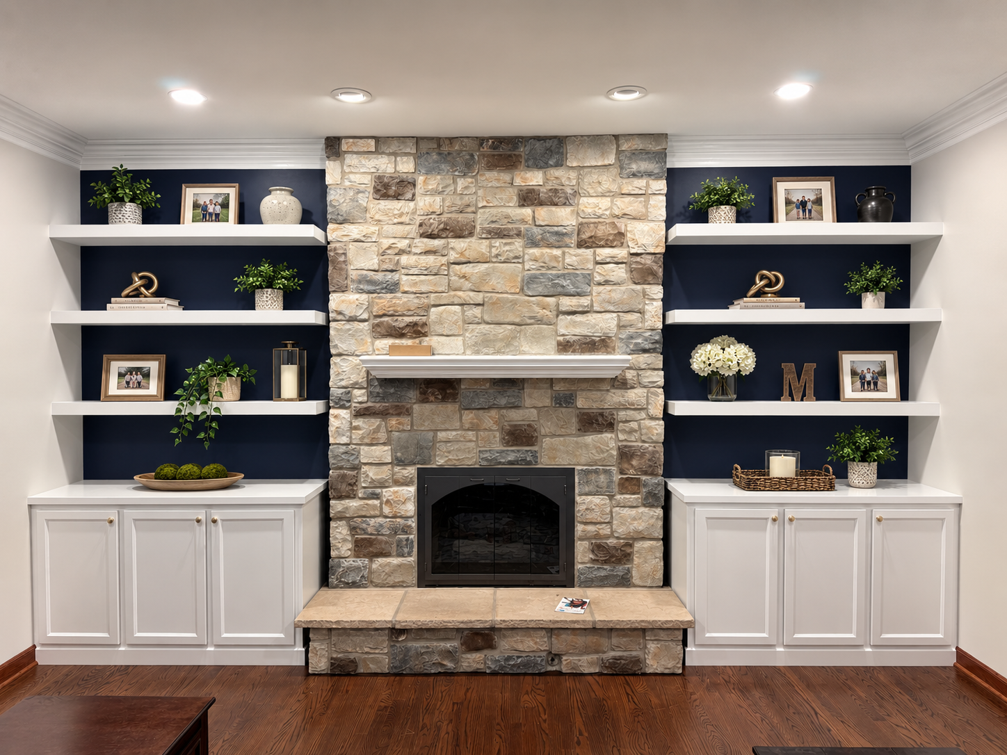 Living room with a stone fireplace, white built-in cabinets, and dark blue accent walls with white floating shelves decorated with framed photos, potted plants, and decorative objects.