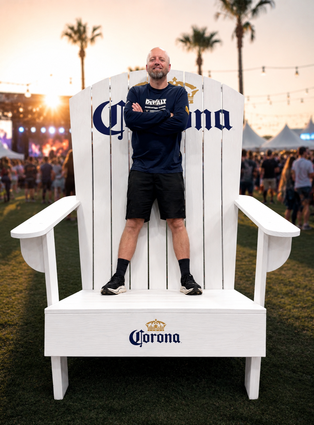 A man standing with arms crossed on a large white Corona beer branded chair at an outdoor event during sunset. There are palm trees and a crowd in the background.