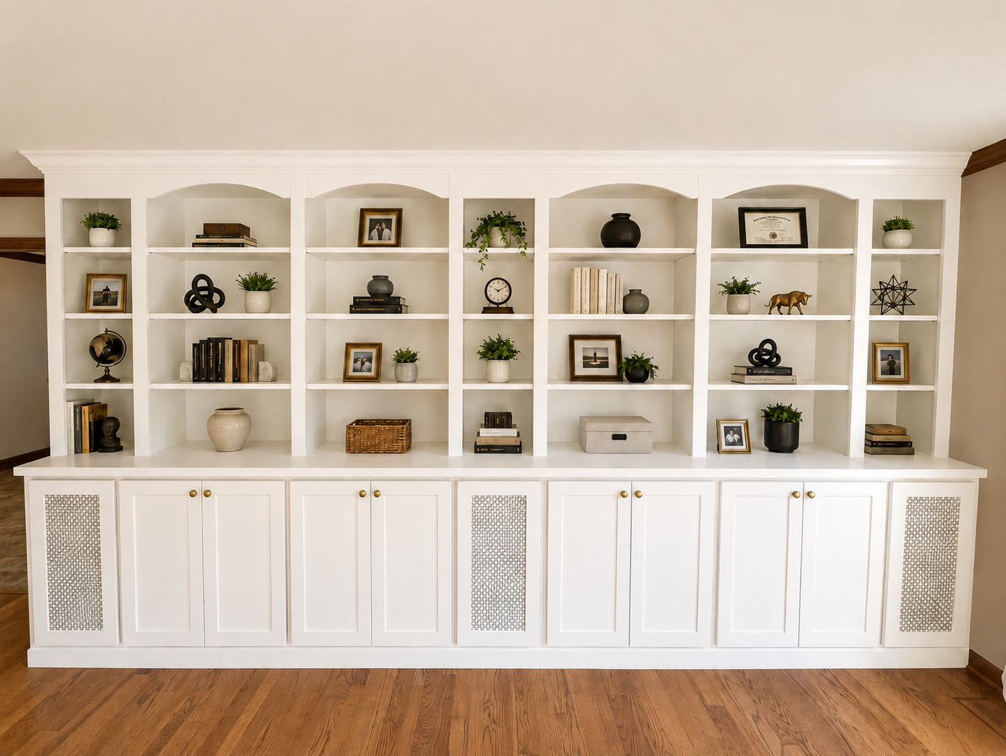 White built-in bookshelf with decorative items, books, framed photos, plants, and a closed cabinet base against a light-colored wall with wooden flooring.