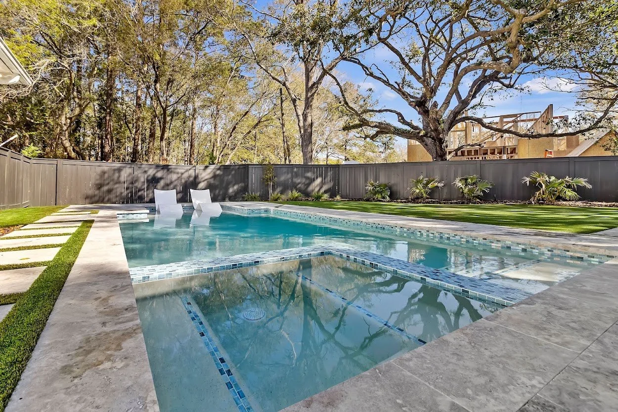Backyard with swimming pool, two white lounge chairs, lush green grass, trees, and a gray fence.
