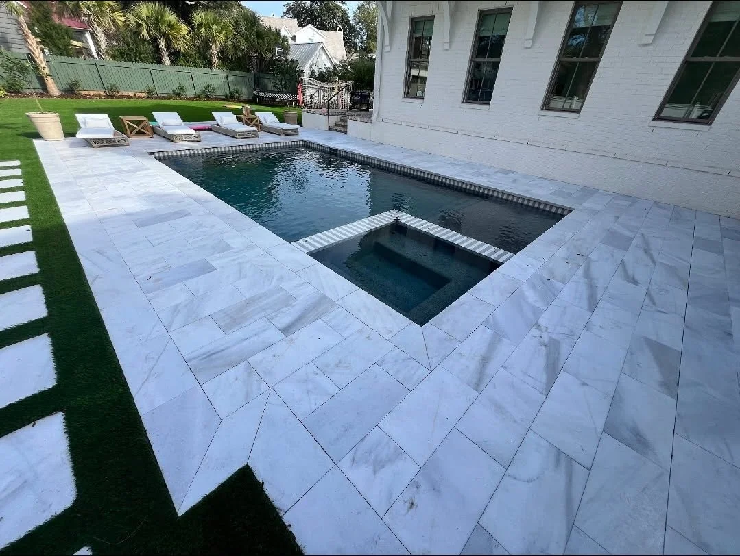 Backyard with a rectangular swimming pool surrounded by white marble tiles, four lounge chairs with cushions, and a green lawn with stepping stones. A white house with multiple windows is visible on the right.