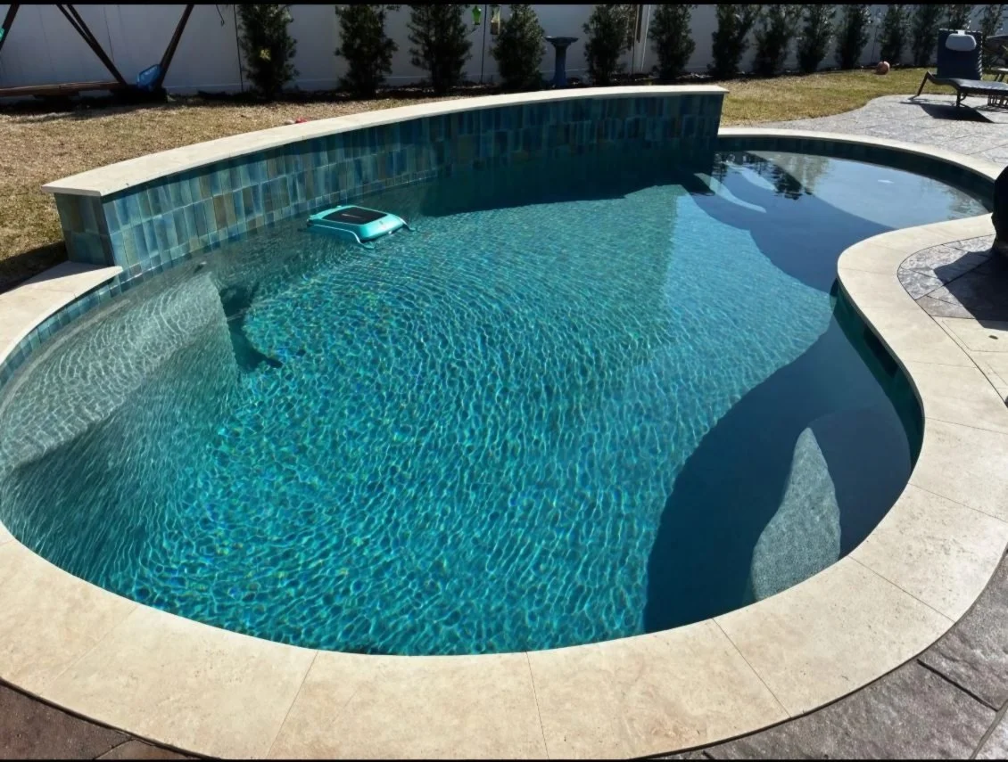 A backyard swimming pool with clear water, a skimmer on the surface, and a stone-tiled edge. There is a patio with chairs and a grassy yard with trees and a fence in the background.